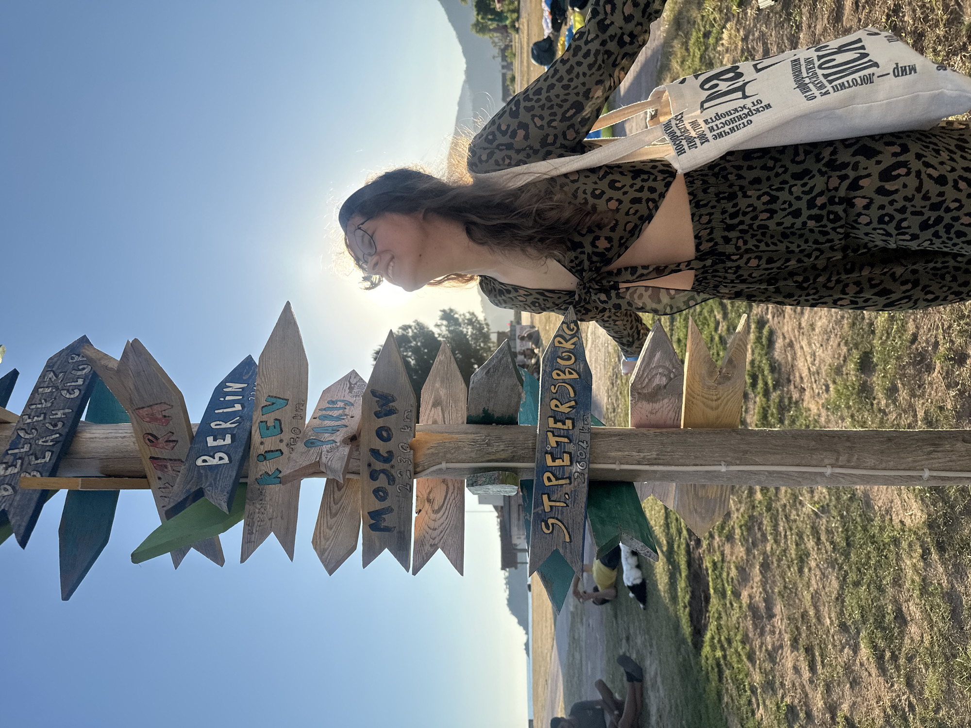 The image shows a young woman standing next to a wooden signpost with various city names painted on the signs, in the city of Fethiye, Turkey. The signs include "Belcekiz Beach Club", "Berlin", "Kiev", "Moscow", and "St. Petersburg". The woman is wearing a leopard-print dress and has a white tote bag slung over her shoulder. The sun is shining brightly, and the sky is blue. There is a grassy field in the background. The text in Russian is present on the tote bag. In the background, people can be seen lying down on the grass.