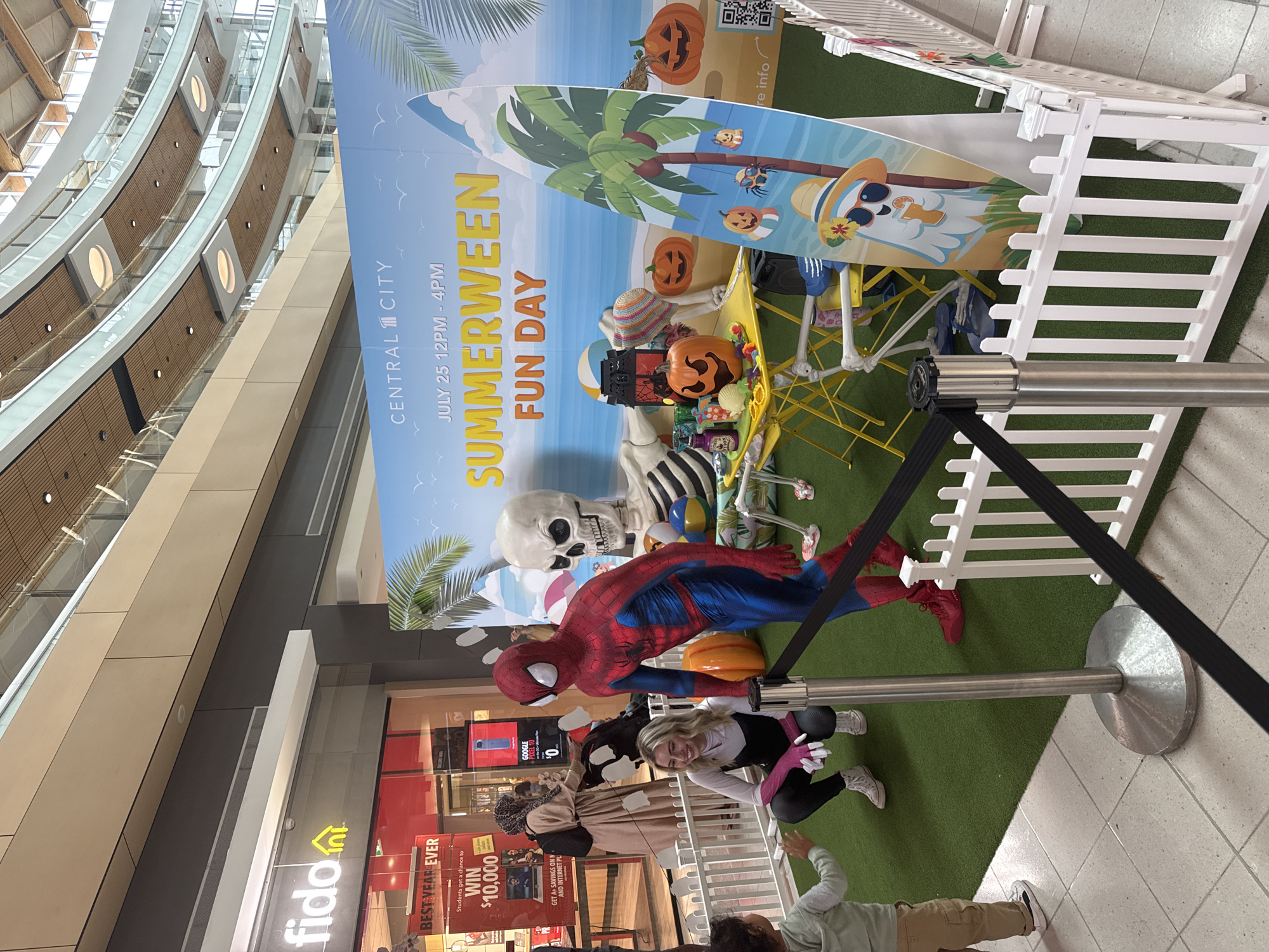 The image depicts a "Summerween Fun Day" event held indoors at Central City in Surrey, British Columbia, Canada. In the foreground, a person dressed as Spider-Man stands near a young child. To the left, a woman dressed as Ghost-Spider crouches down, interacting with the child. Behind them, a large skeleton prop is seated at a yellow table laden with Halloween-themed decorations, including pumpkins and lanterns. A surfboard-shaped cutout featuring a cartoon ghost enjoying a drink on a beach is also visible. The event appears to be set up on a section of artificial turf, enclosed by a white picket fence and stanchions with black tape. In the background, a Fido store is visible, along with escalators and multi-level architecture characteristic of a shopping mall.