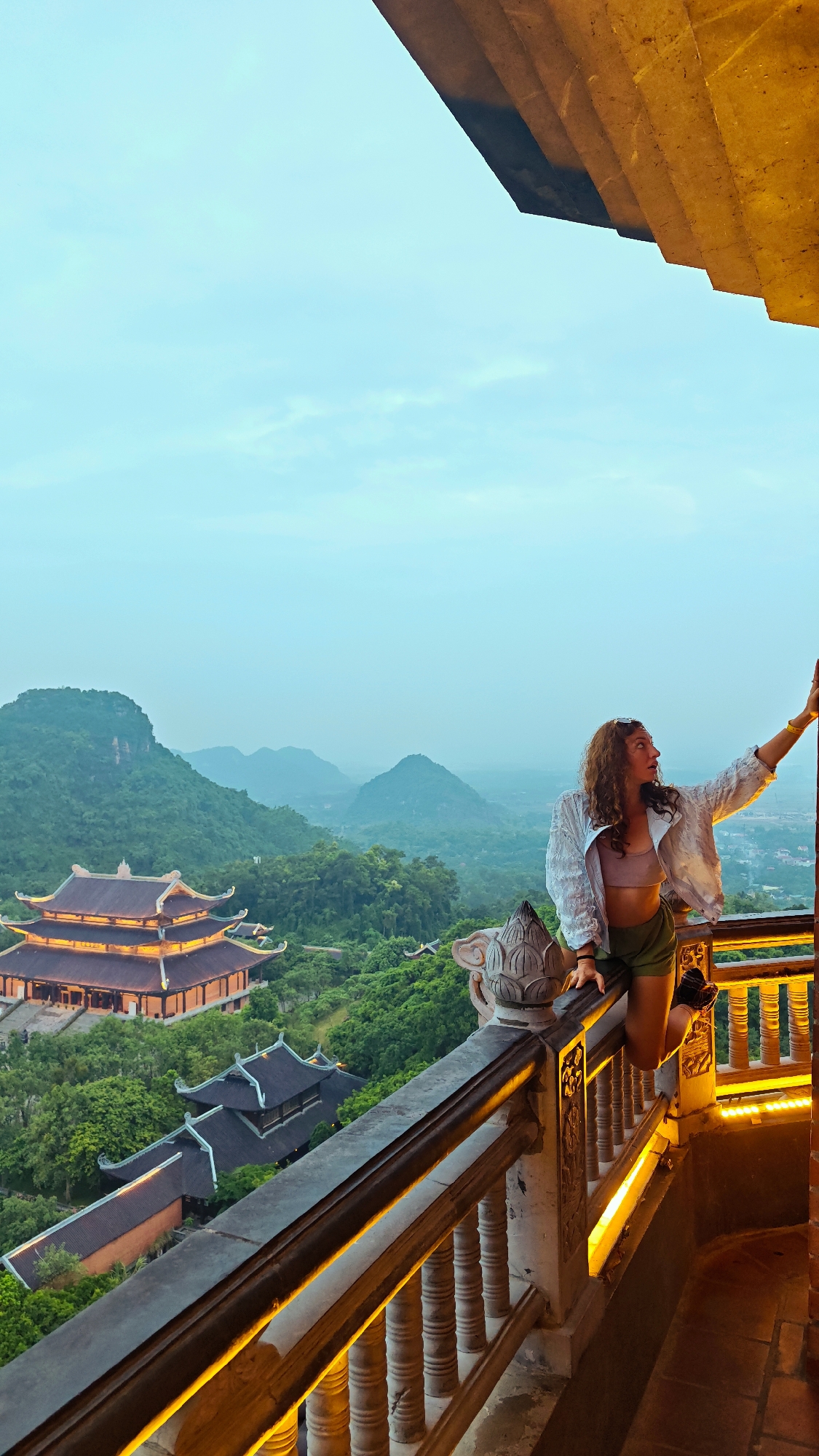 A young woman with curly brown hair stands on a balcony overlooking a lush green valley with traditional Vietnamese architecture. She is wearing a white open shirt over a pink crop top and green shorts. Her right arm is extended, resting on a railing that features ornate carvings and is illuminated by warm yellow lights. In the background, several pagoda-style buildings with orange roofs are nestled amongst dense trees. Rolling green hills and mountains form the distant landscape under a pale blue sky. The woman is looking away from the camera towards the scenic view.