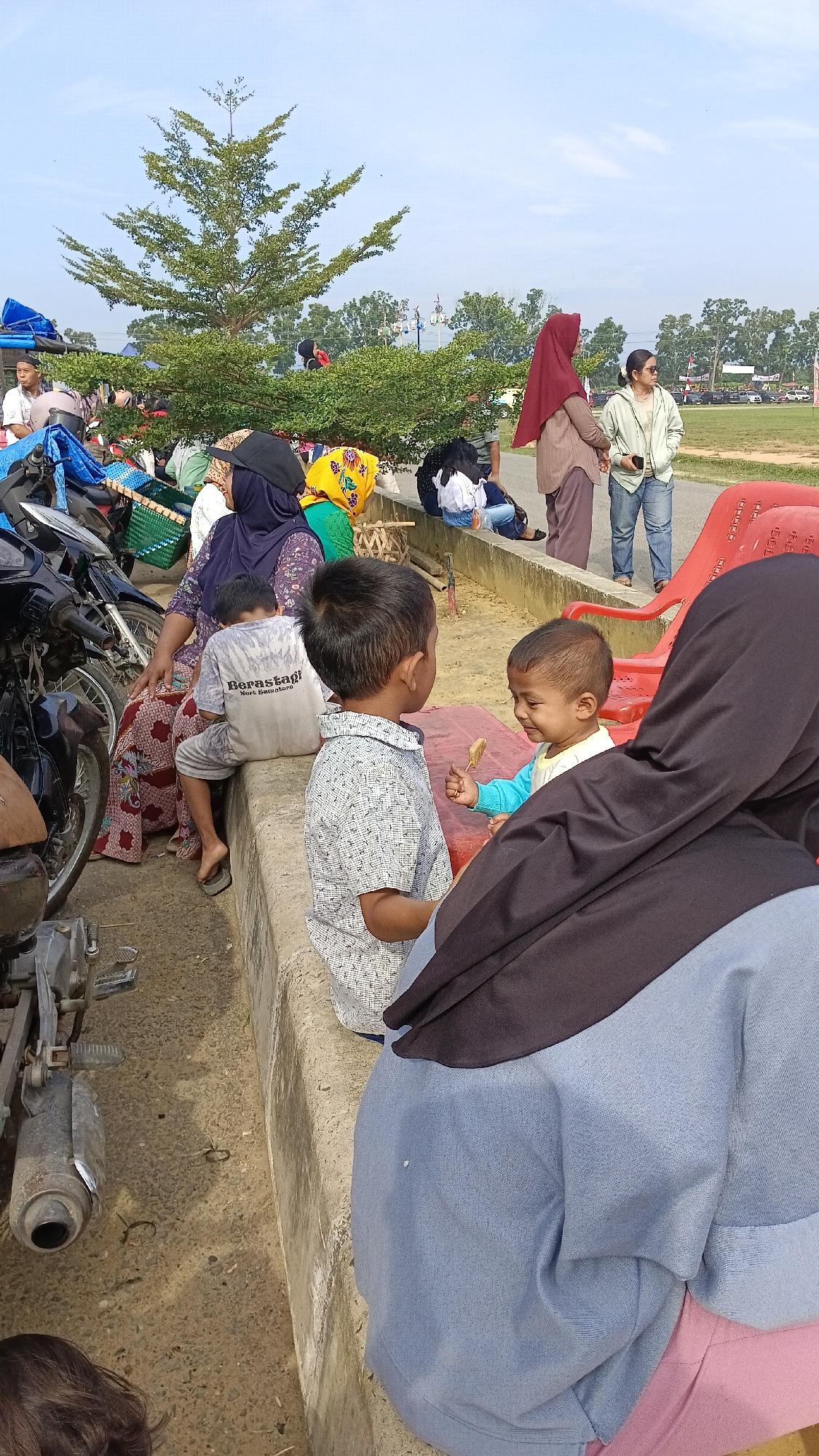In Aek Kanopan, Indonesia, on a sunny day, a group of people gather outdoors. In the foreground, a woman wearing a black hijab and a blue jacket is seated, with her back mostly to the camera. To her right, a young child in a blue and white shirt is being offered a snack by an older boy in a patterned shirt. They are seated on a low concrete ledge. Behind them, a woman in a floral patterned dress and a purple hijab is sitting, holding a bag. To her left, a motorcycle is parked. In the middle ground, other people are seated or standing, some wearing hijabs of various colors. A large green tree with sparse leaves stands prominently in the background. Further beyond, there are more trees and a grassy field, suggesting an open area or park. The sky is clear and blue.