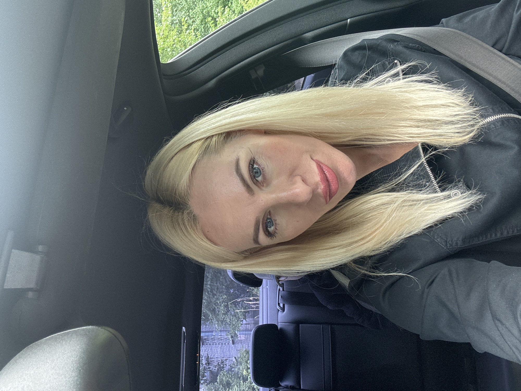A young, blonde woman with blue eyes and rosy lips is taking a selfie inside a car. She is wearing a black jacket with a zipper. The interior of the car is dark, with black leather seats visible in the background. The car appears to be moving, as indicated by the blurred greenery outside the window, suggesting a daytime setting on a clear day. The woman's pose and direct gaze into the camera convey a sense of confidence and engagement. The overall mood is casual and relaxed, typical of a personal snapshot. There is no visible text in the image. The location context of Saint Petersburg, Russia, is not directly evident from the visual information in the image itself.