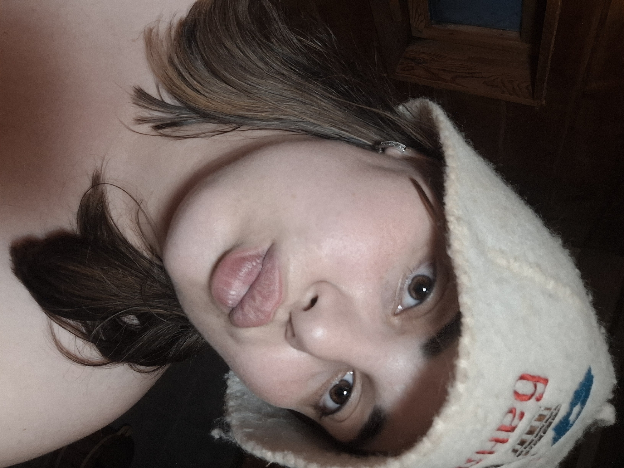 A close-up, angled portrait of a woman wearing a white felt hat with Cyrillic script in red and blue. Her hair is dark brown with highlights. Her expression includes slightly puckered lips. The background is a dark, wooden wall. The location may be Амзя, Russia, based on the Cyrillic script on the hat.