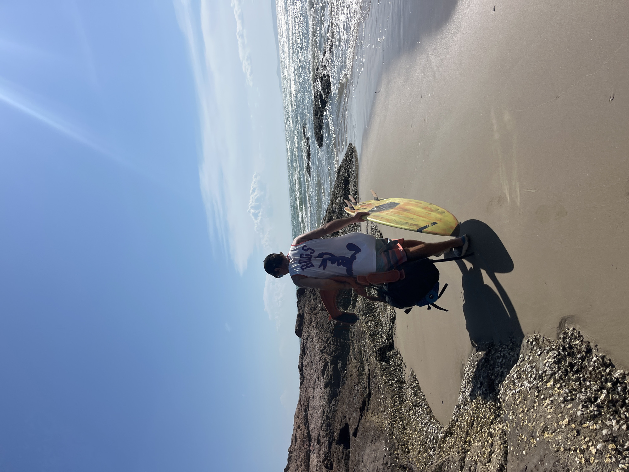 A person wearing a white tank top with a purple "Bugs" graphic and red and white patterned shorts walks along a sandy beach. They are carrying a black backpack and a yellow and orange surfboard. The person is walking away from the camera towards a body of water with choppy waves. To the left of the person is a rocky area covered in shells. The sky is a clear blue with a few white clouds. The scene appears to be a coastal environment, potentially in a tourist area given the presence of surfboards. This specific location in Mũi Né, Vietnam, is characterized by its scenic beaches, rocky outcrops, and the popular water sport activities.