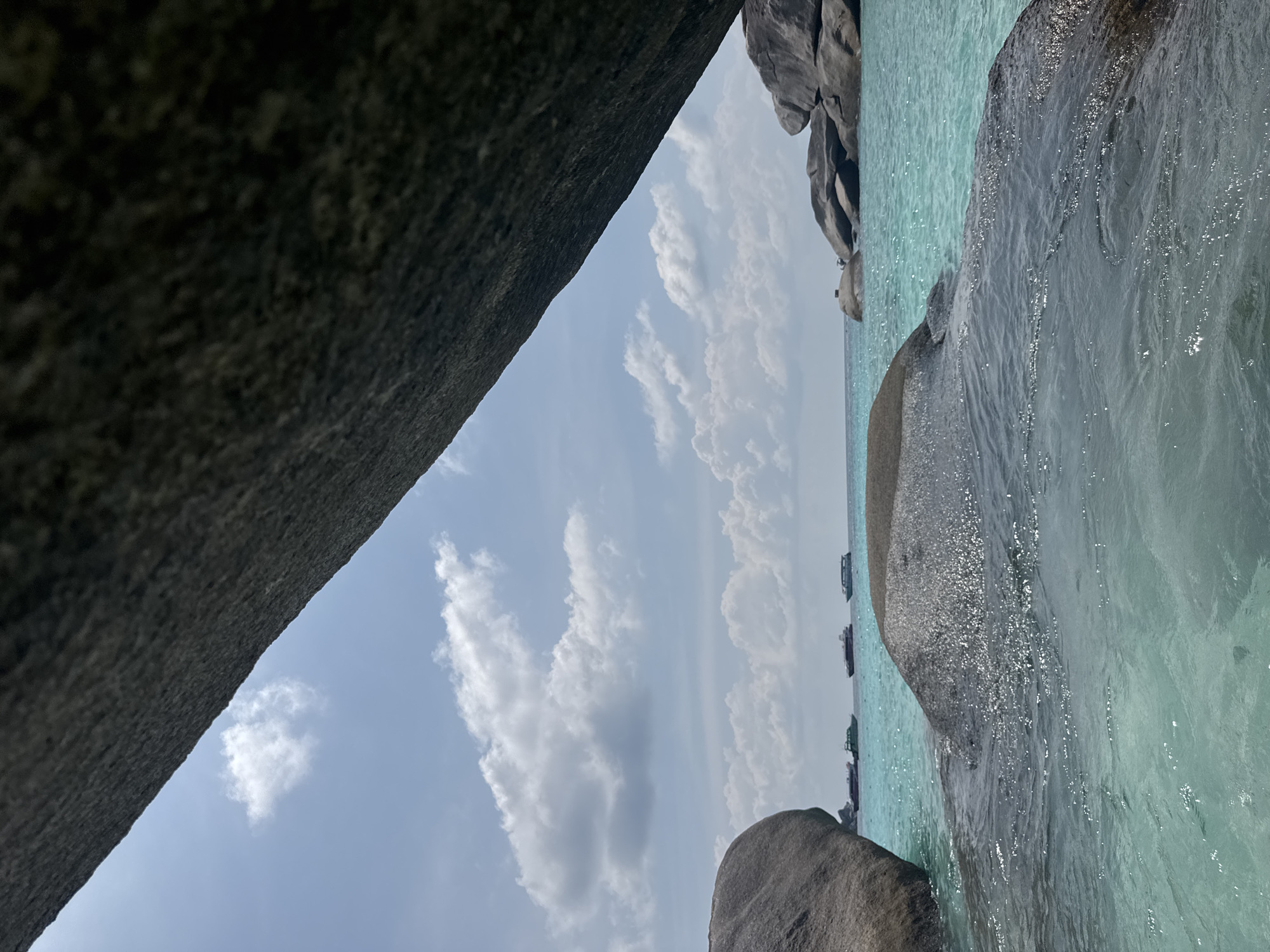 The image captures a view from what appears to be a rocky outcrop in Lam Kaen, Thailand. The foreground is dominated by large, dark gray rocks forming a natural frame. Between the rocks is a view of the turquoise sea and a partly cloudy sky. Several boats can be seen on the horizon. Sunlight glints off the water, creating a shimmering effect.