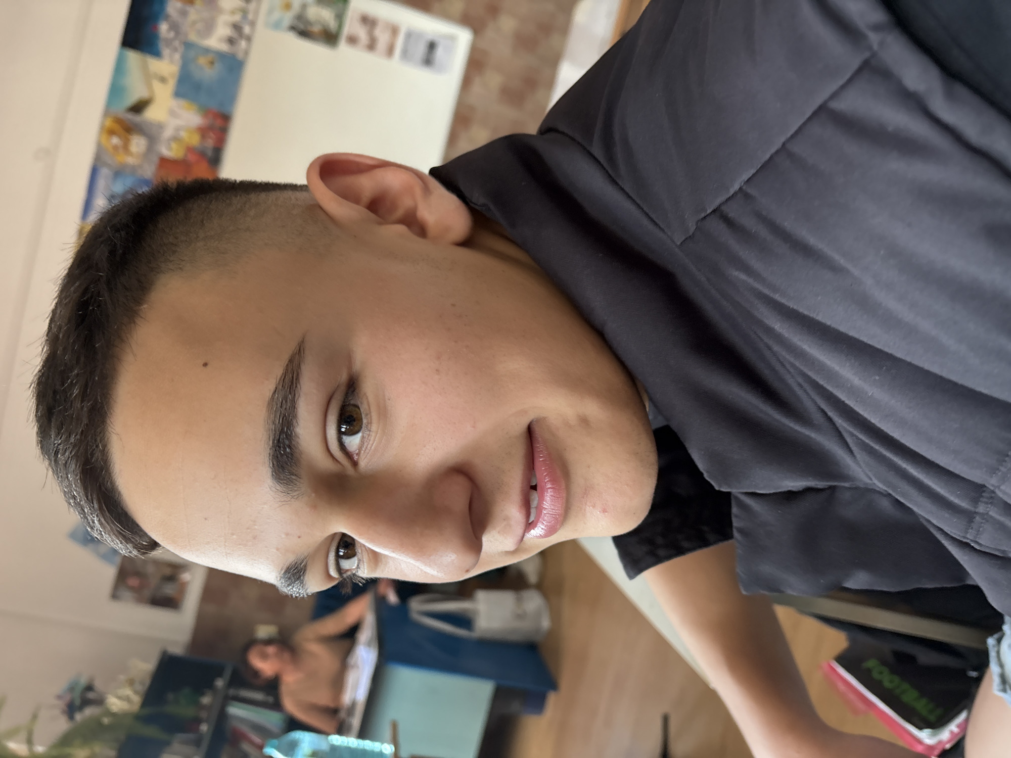 A close-up, indoor shot focuses on a person with short, dark hair and light skin, their face angled towards the camera. They wear a dark-colored shirt or jacket. The background is somewhat blurry, showing what appears to be a room with artwork on the walls and a person visible in the distance. A hint of furniture is seen. There is no information or evidence in the image to support a location in Panagyurishte, Bulgaria.
