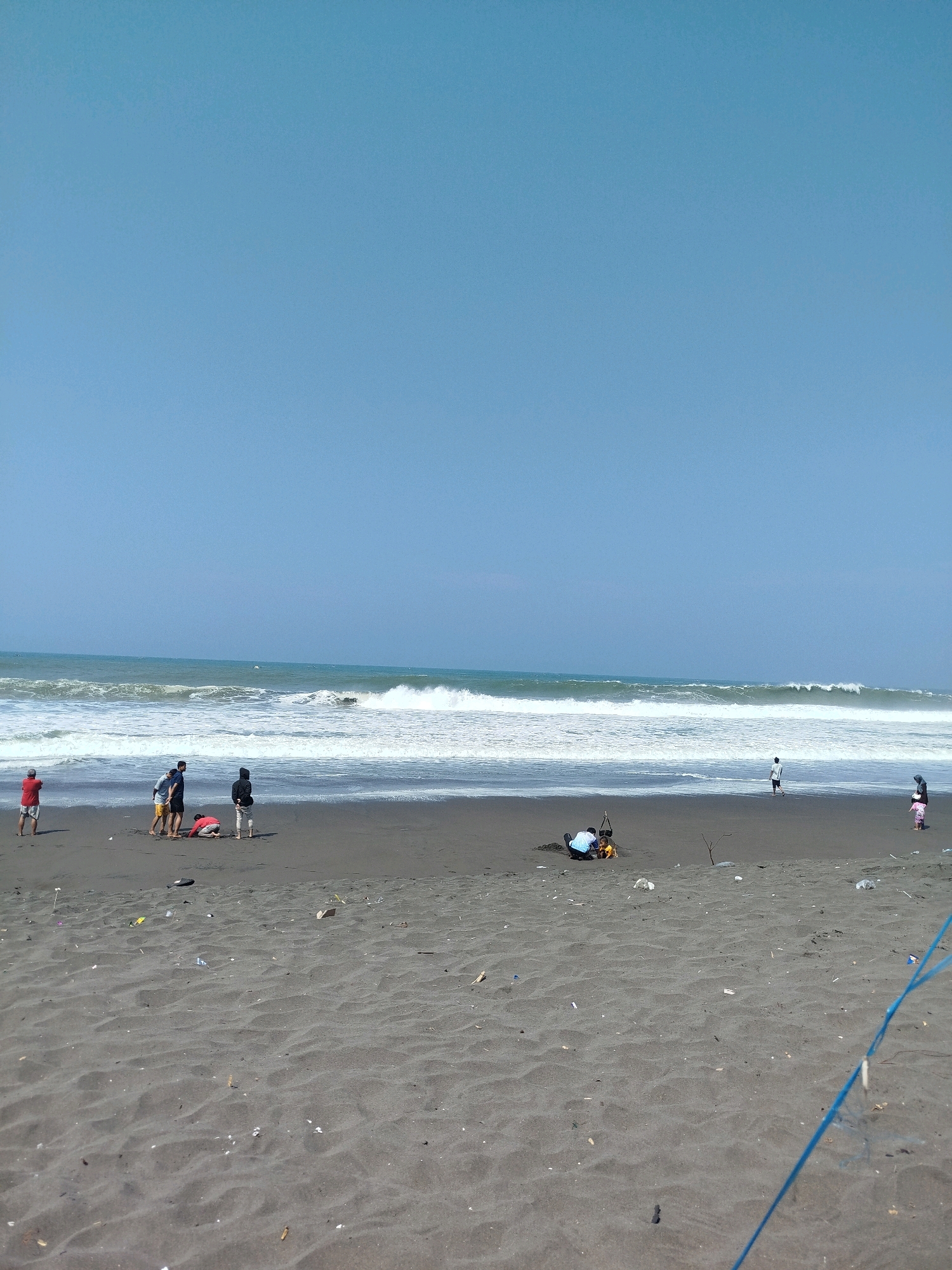 A wide, low-angle shot captures a sandy beach under a clear blue sky. The foreground is dominated by dark, rippled sand, dotted with scattered pieces of litter, including plastic wrappers and debris. In the mid-ground, a group of people are gathered near the water's edge. A group of four individuals are clustered together, with one person in a red shirt standing slightly apart. Another group of two individuals, one in dark clothing and another in a hooded jacket, are also visible. In the distance, waves are crashing on the shore, creating white foam. Further out, the ocean stretches to the horizon under the bright sky. On the right side of the frame, a lone individual in white walks along the shoreline, and a woman in a pink skirt and headscarf stands near the water. The scene appears to be a casual gathering on a beach in Kenoyajaya, Indonesia.