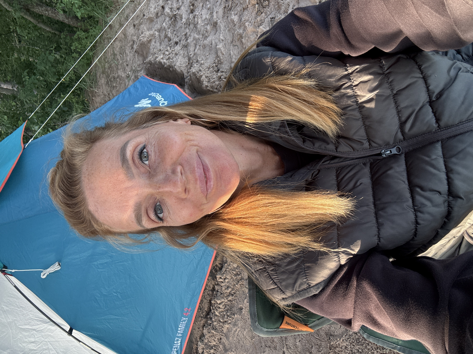 In Shepelevo, Russia, a woman is seated in a folding camping chair next to a blue and white tent. She is wearing a dark gray puffer vest over a dark purple hoodie. Her light brown hair is shoulder-length, and she is looking at the camera with a slight smile. The tent is set up on a sandy surface, with trees visible in the background. The sun is casting warm light on the scene, highlighting the woman's face and hair.