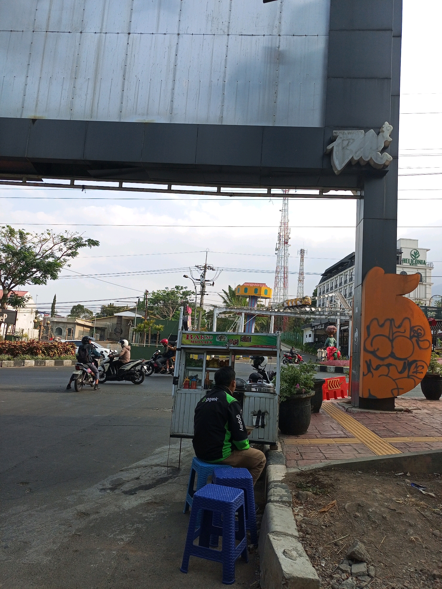 The image captures a street scene in Batu, Indonesia, likely near a commercial area. In the foreground, a person wearing a black and green jacket with "gojek" branding sits on a blue plastic stool facing away from the camera, near a food cart. The food cart has signs in Indonesian, one reading "BARSO MUSTAFA". To the left, several motorcycles are visible on the road, with riders wearing helmets. One motorcycle is parked near the food cart. In the mid-ground, a small amusement park ride with a yellow train is operational, with a yellow structure above it. In the background, there are buildings, including one with "HELICONIA HOUSE" written on it, and several communication towers. The sky is partly cloudy. The overall environment suggests a bustling urban setting with various activities taking place.