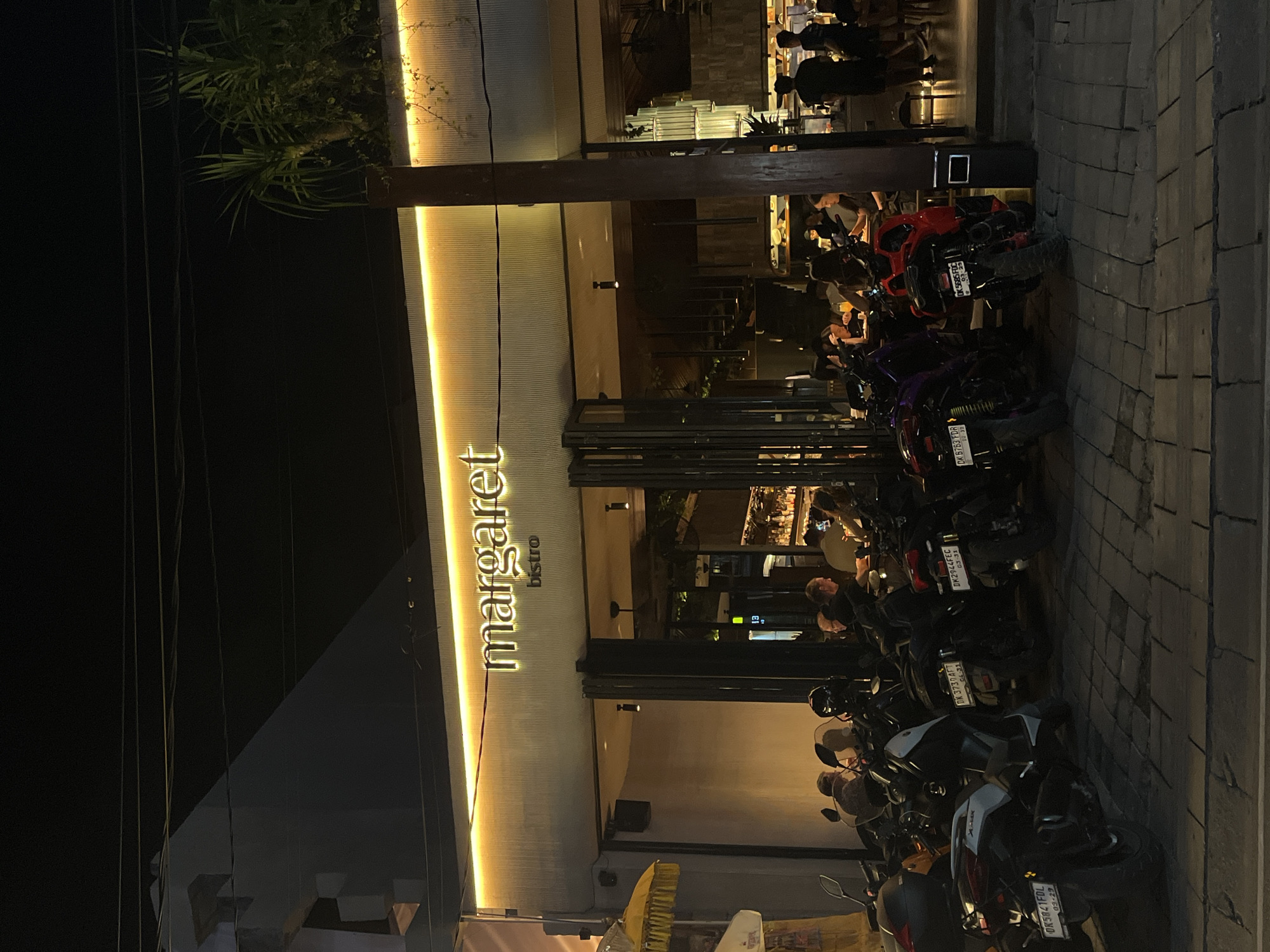 The image depicts the exterior of "Margaret Bistro" in North Kuta, Indonesia, at night. A neon sign with the bistro's name is illuminated above the entrance. The entrance features large glass doors that are partially open, revealing patrons seated at tables inside. Several motorbikes are parked in front of the establishment on a cobblestone pavement. The scene is dimly lit, with the primary illumination coming from the bistro's sign and interior lighting.