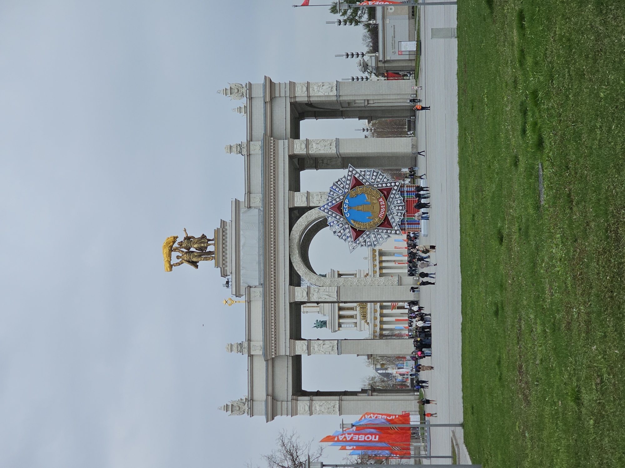 This image displays the grand Main Entrance Arch of VDNKh (Exhibition of Achievements of National Economy) in Moscow, Russia, on an overcast day.
The central subject is a monumental, light-colored triumphal arch, featuring multiple tiers, classical columns, pilasters, and intricate decorative carvings. Perched atop the left side of the arch are golden statues of two figures, possibly workers or farmers, collectively holding a large sheaf of wheat, an iconic symbol of the location.
In front of the arch, a very large, prominent star-shaped emblem is displayed. This emblem has a central blue medallion depicting two golden structures, likely Moscow Kremlin towers, and is surrounded by red and white sections. Clearly visible Cyrillic text on the emblem reads "CCCP" (USSR) and "ПОБЕДА" (Victory), strongly suggesting a commemoration related to Victory Day.
A wide, paved walkway stretches across the foreground and extends under the arch, where numerous people are seen walking and observing the surroundings. To the right of the walkway, a broad expanse of vibrant green grass fills a significant portion of the frame. In the distance, through the archways, glimpses of other buildings typical of the VDNKh complex can be seen. On the far left, several red and blue banners are visible, also bearing the Cyrillic text "ПОБЕДА!" (Victory!).
The scene captures a public space bustling with visitors enjoying the architectural grandeur. The soft, diffused light from the cloudy sky indicates daytime. The presence of the "Victory" symbols and Soviet-era architecture points towards a site of historical and national significance, likely related to a commemorative event in Moscow.