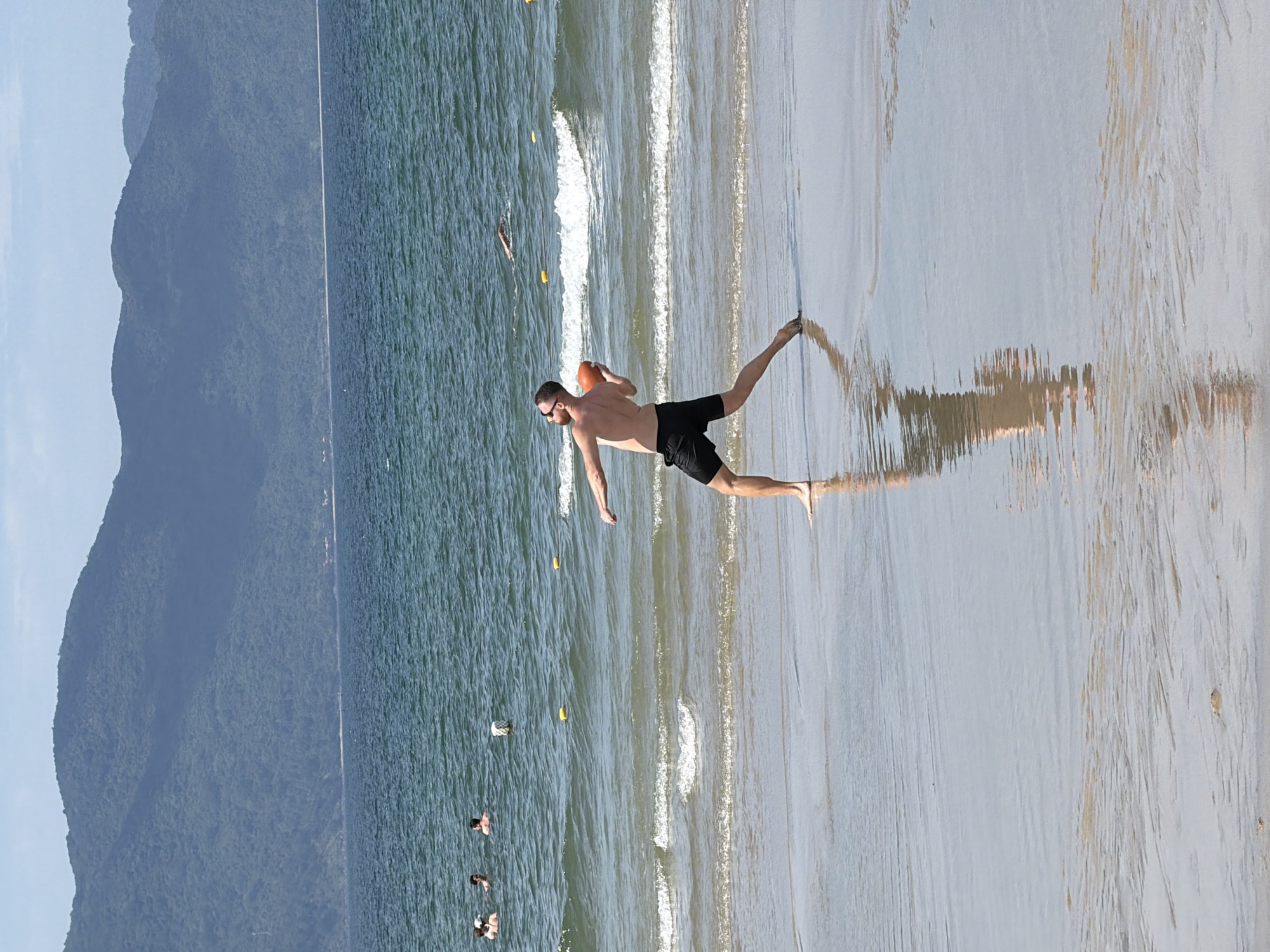 A man is playing with a ball on the beach in Da Nang, Vietnam. He is wearing black shorts and sunglasses, and he is throwing an orange ball. The sea is blue and there are waves. In the background, there are mountains. There are a few other people in the water. The sand is wet and there are reflections of the man and the sky on the wet sand.