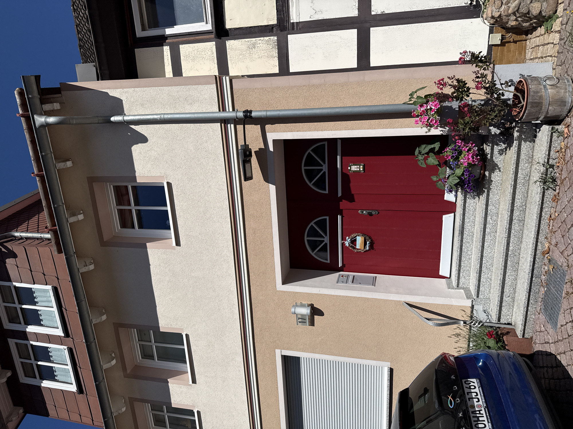 The image shows the entrance to a multi-story building, likely a residential property, located at Marktstraße 17 in Bad Sachsa, Germany. The building has a light beige stucco facade on the lower two stories and a darker reddish-brown tiled upper story. Several windows with white frames are visible on all levels, some with white curtains. A large red double door with a semicircular fanlight is the central feature of the ground floor entrance, flanked by a mailbox and a doorbell. A wreath with a blue ribbon is hanging on the left door. A metal handrail curves up a set of granite stairs leading to the entrance.
To the left of the entrance, a blue car with a license plate reading "OHQ JJ 69" is parked on a cobblestone driveway. A metallic garage door with horizontal slats is also visible on this side of the building. A silver downspout runs vertically along the right side of the entrance. Potted plants with pink and purple flowers are placed on the stairs and a wooden barrel planter sits near the entrance, containing a small tree with pink flowers.
To the right of the entrance, the building transitions to a different architectural style, featuring timber framing with cream-colored panels and dark brown beams. A small stone wall forms the base of this section of the building. The sky is clear and blue. The overall scene depicts a well-maintained residential property on a sunny day.