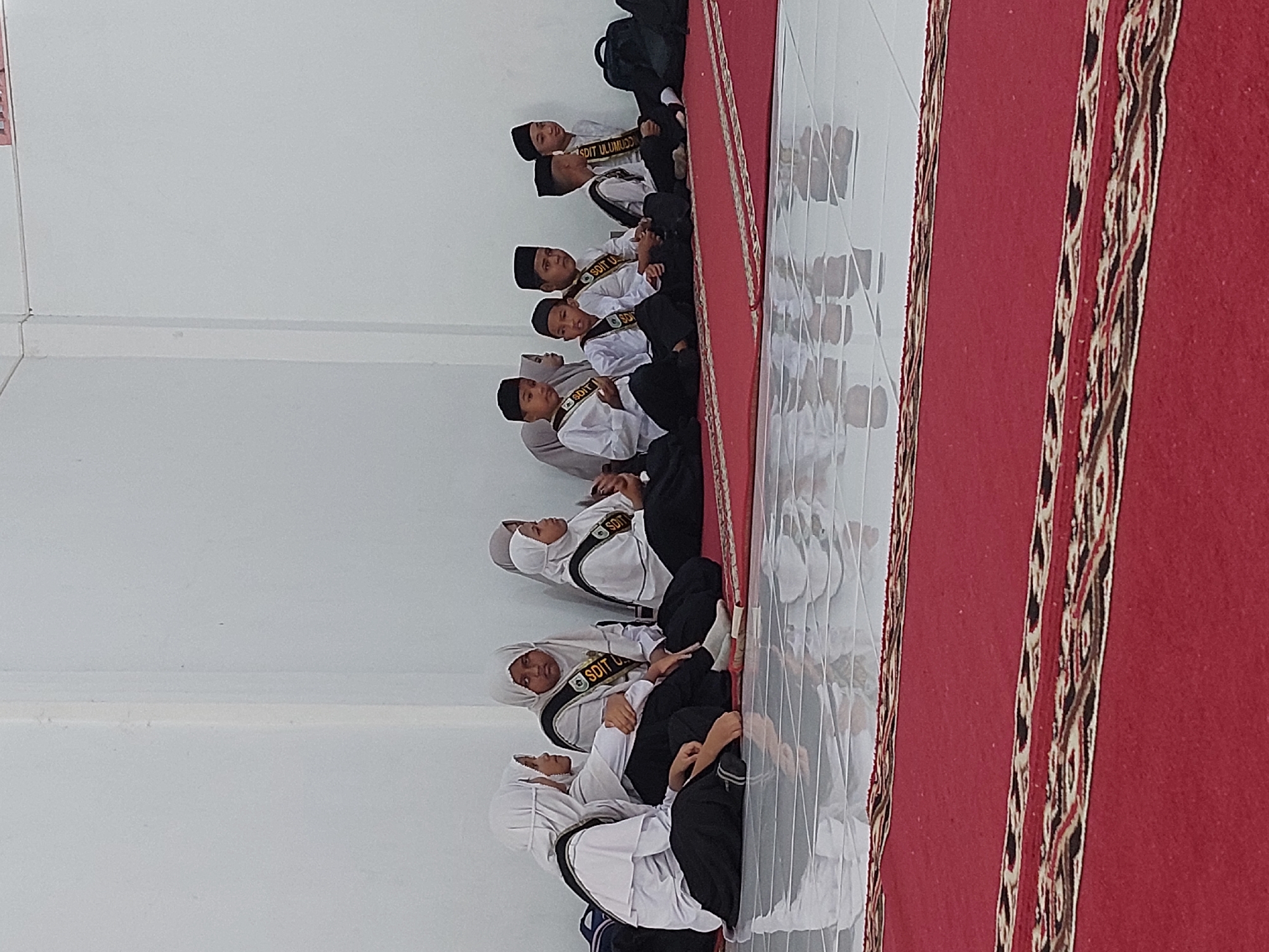 A ground-level, medium-distance shot shows a group of Indonesian students sitting in rows on a red rug inside what appears to be a mosque or a school classroom. The location is Lhokseumawe, Indonesia, and the students are wearing white shirts, black pants, and black hats, with some female students wearing white headscarves (hijabs). They also have black and gold sashes with the words "SDIT ULUUMUDDIIN" printed on them. They are sitting facing an unseen point in the room. The room has white walls and a light-colored tiled floor. The photograph is oriented sideways.