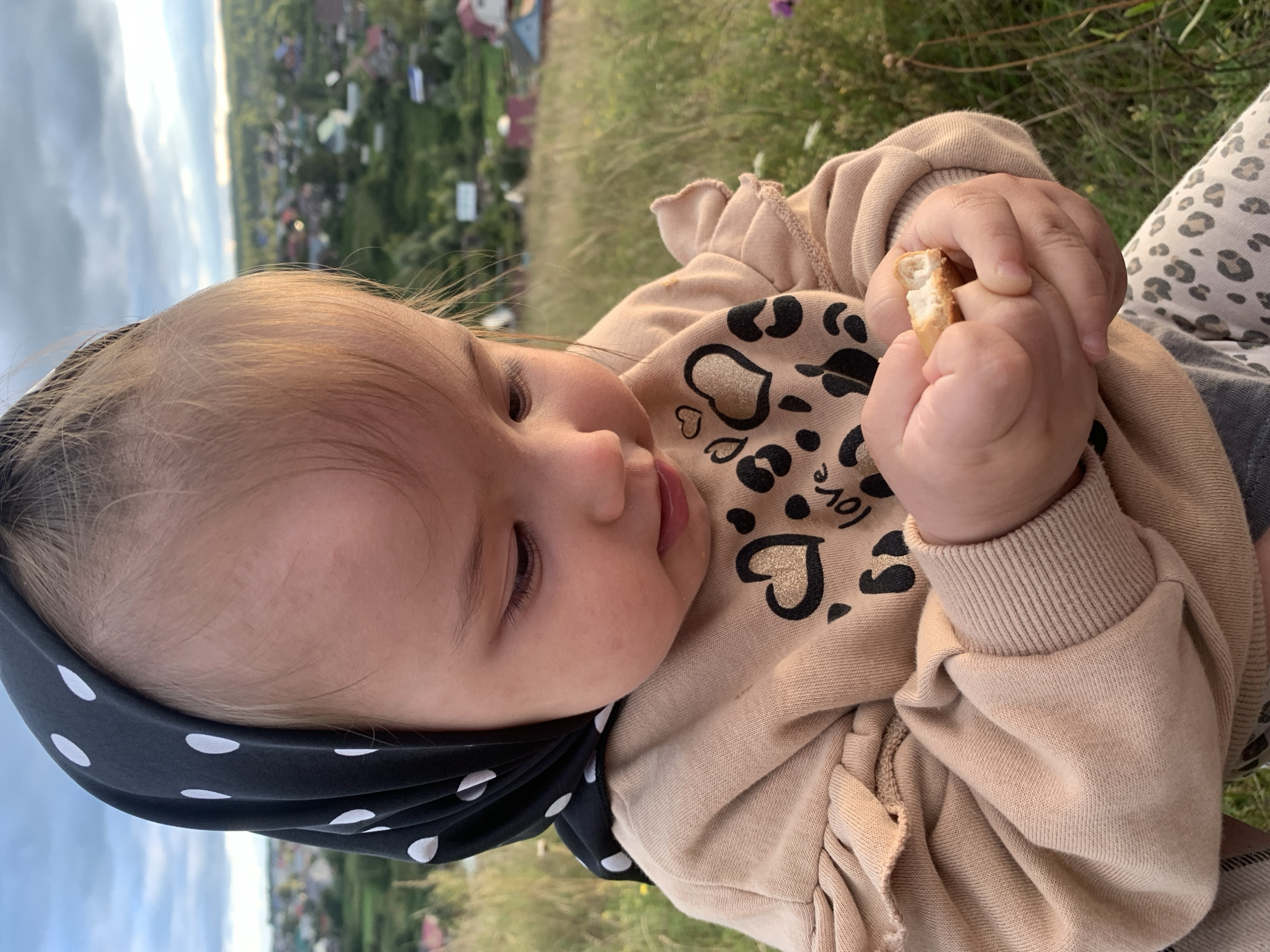 A close-up, eye-level shot shows a young Caucasian baby girl wearing a black and white polka dot headscarf and a tan sweatshirt with a leopard print and heart design. She is holding a small piece of cracker in her hands and looking down at it. The background is a blurred rural landscape with houses and trees, suggesting the location is in or near Olkhovka, Russia. The lighting indicates it is late afternoon or early evening, with soft sunlight casting a warm glow. The grass and vegetation in the foreground and background are green and natural.