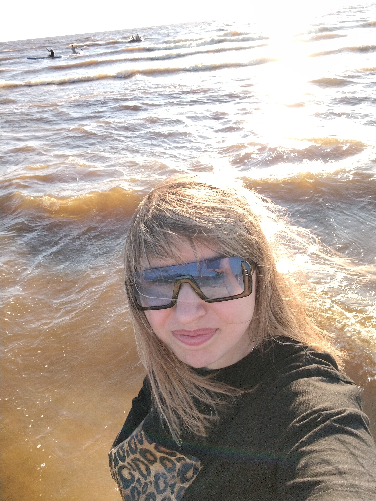 In Ivanovskoe, Russia, a selfie captures a woman in large, dark sunglasses, her blonde hair windswept. She is wearing a black long-sleeved shirt and her arm is adorned with a leopard-print sleeve. She is positioned in the foreground, with the shimmering water of a lake or sea stretching out behind her. The water is choppy with gentle waves, reflecting the bright, setting sun which casts a warm glow and lens flare across the scene. In the distance, silhouetted figures can be seen on paddleboards, one of which appears to have a bird flying nearby. The overall impression is of a peaceful, outdoor recreational moment by the water.
