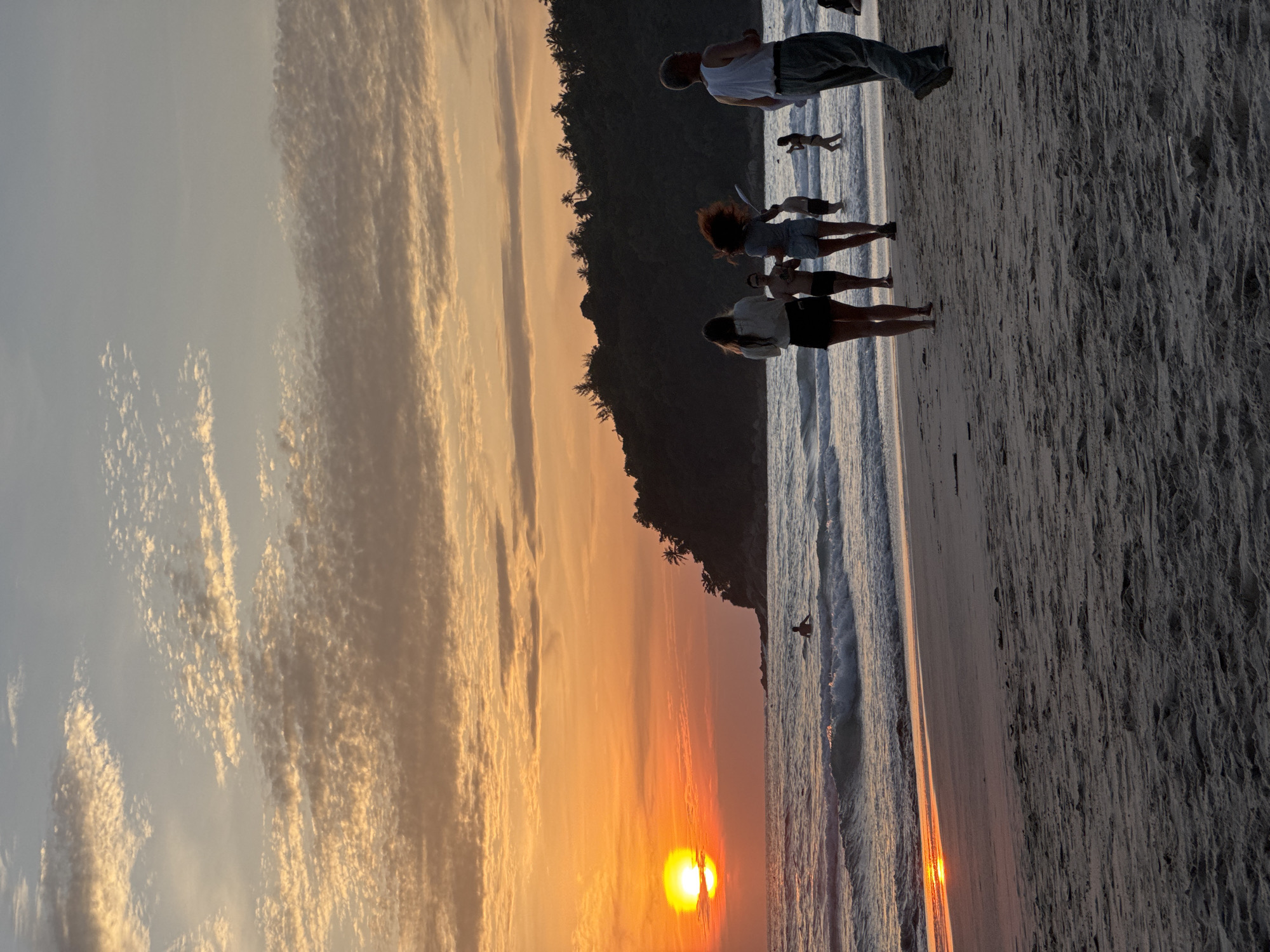 The image depicts a sunset on Karon Beach in Karon, Thailand. The sun is a bright orange orb positioned low on the horizon, casting a warm glow across the sky, which is filled with wispy clouds tinged with orange and yellow hues. Gentle waves are breaking on the sandy shore. Several people are visible on the beach and in the water. In the foreground, on the sand, are three individuals walking towards the sea, and one person walking away from the sea. Further down the beach, several other people are scattered, some in the shallow water. In the distance, a tree-covered hill forms the backdrop against the vibrant sky.