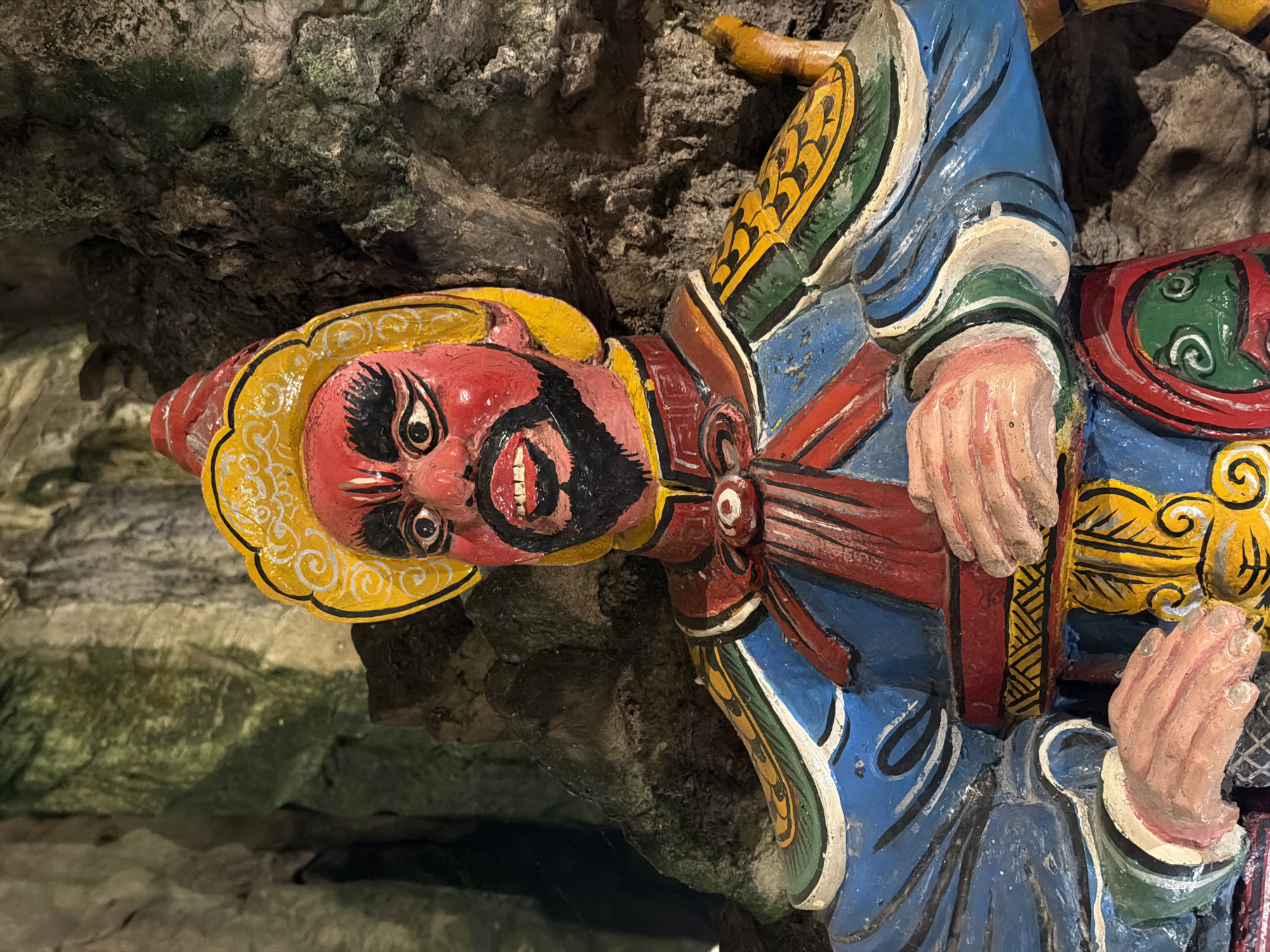 This media file showcases a vibrantly painted, sculpted figure of a deity or mythological character, set against a rugged, natural cave wall. The image is presented sideways, suggesting the figure is mounted vertically on a wall or the photograph was taken with a rotated camera.
The figure’s face is a striking red, framed by a prominent black beard and mustache. It features wide, intense eyes with black pupils and white outlines, and a broad, open mouth revealing white teeth, conveying a fierce or powerful expression. A distinctive yellow headpiece, adorned with intricate, swirling gold patterns, crowns its head.
The body of the figure is arrayed in elaborate robes, predominantly blue, accented with sections of red, green, and yellow, detailed with white and black patterns. Its hands, depicted in a flesh tone, are visible; one appears to grasp the hilt of a sword or staff, while the other is clenched, possibly in a gesture of power or readiness.
The background consists of rough, uneven rock formations in shades of grey, green, and brown, indicating a natural cave or grotto environment. The sculpture itself appears to be either carved into or affixed to this rocky surface. The vivid colors and detailed craftsmanship suggest a significant cultural or religious artifact, likely found in a temple or sacred site in Đà Nẵng, Vietnam. No visible text is present.