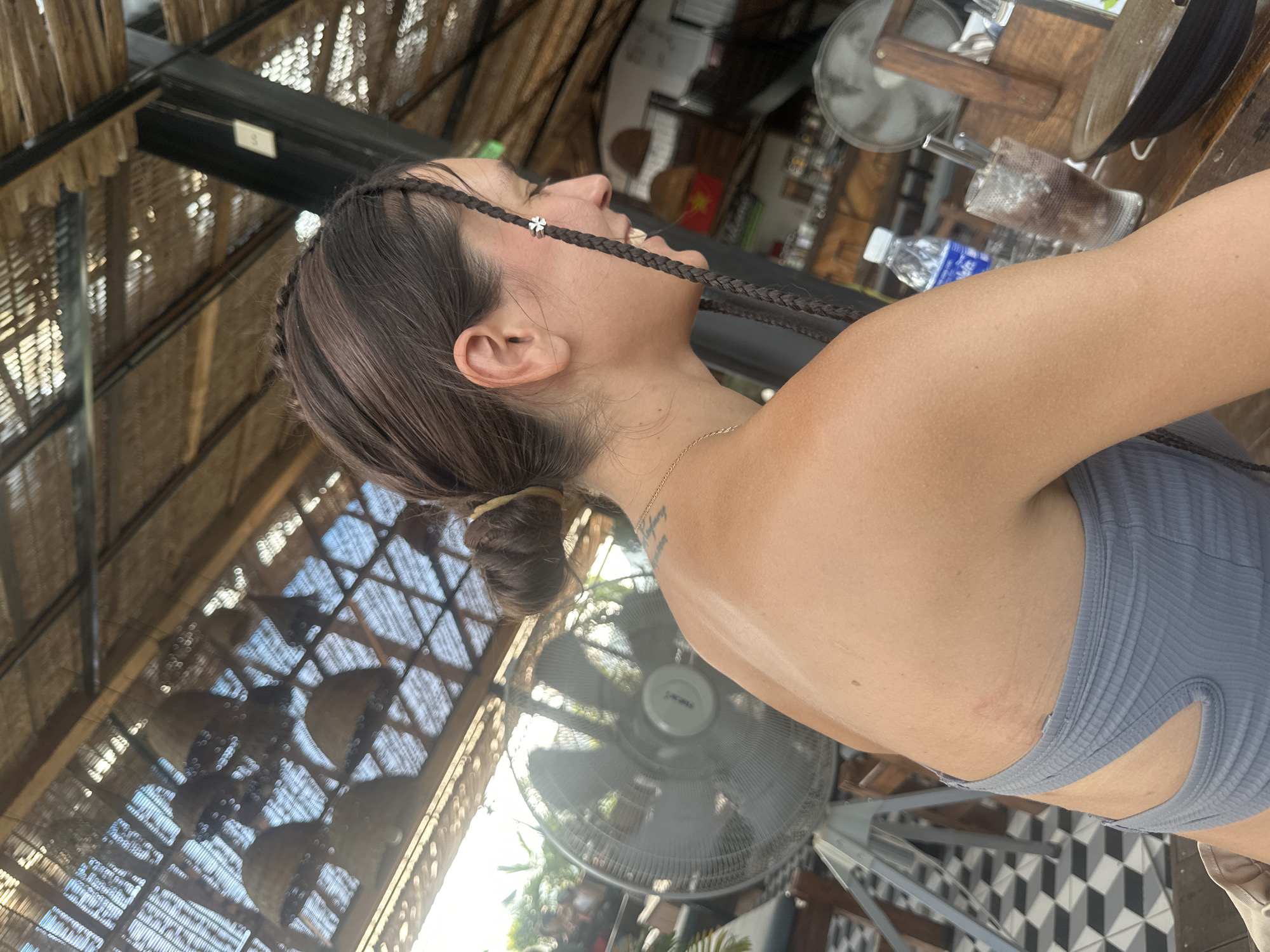 The image presents a profile view of a person, likely female, captured from the right side, looking upwards and slightly to the right. She has dark hair, styled with two thin braids framing her face, one adorned with a small silver clover-shaped charm. Her hair is tied in a low bun at the back. Her mouth is slightly open, revealing teeth, indicating a smile or laughter. A thin gold chain necklace is visible around her neck, and a faint tattoo is on her upper back. She is wearing a light gray, ribbed top with cut-out details on the side.
The setting appears to be an open-air establishment, possibly a cafe or restaurant, characterized by a rustic, woven, or thatched ceiling structure that allows light to filter through. Several items are visible in the background: a large standing fan (brand "Senko" visible), a tabletop with a glass containing ice and a dark drink, and a plastic "AQUAFINA" water bottle. The floor features a distinct black and white geometric tiled pattern. In the blurred background, a red object with a yellow star is visible, consistent with the flag of Vietnam, confirming the location context of Hà Giang 1 Ward, Vietnam. The scene conveys a relaxed and pleasant moment, likely during the day in a warm environment.