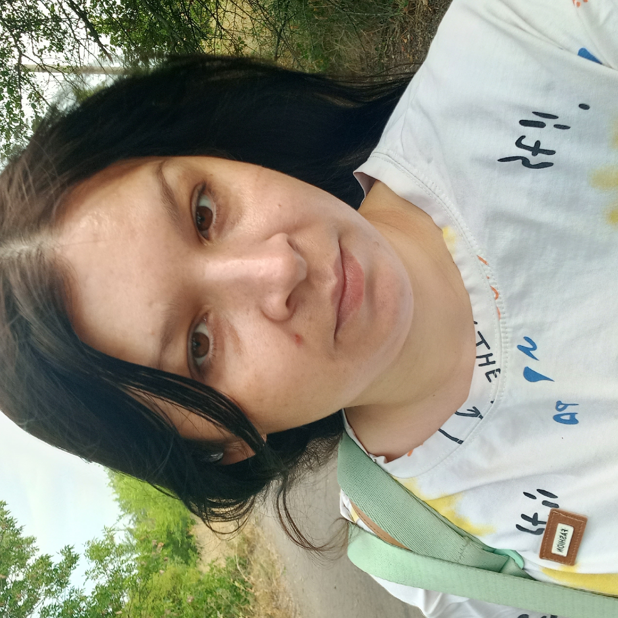 A close-up selfie shows a woman with long, dark hair and brown eyes, facing forward. She has a fair complexion with a small red mark on her cheek. She is wearing a white t-shirt with abstract designs and text, and a light green strap from a bag is visible over her shoulder. The background suggests an outdoor setting, possibly a rural road with trees and vegetation on either side, consistent with a location in or near Neopalymivka, Ukraine. The lighting is natural, indicating it was taken during the day.