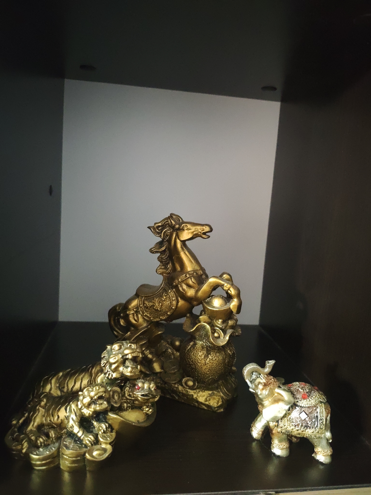 The image shows three golden figurines placed on a dark brown surface, likely a shelf, within a cabinet. The back of the cabinet is divided into a light gray panel on the left and a dark brown panel on the right.
The central figurine is a horse rearing up on its hind legs, appearing to stand on a large golden pot. To the left is a tiger figurine, which appears to be a group of tigers. To the right is a small elephant figurine adorned with colorful decorations. The location of the scene is Unknown, Unknown.