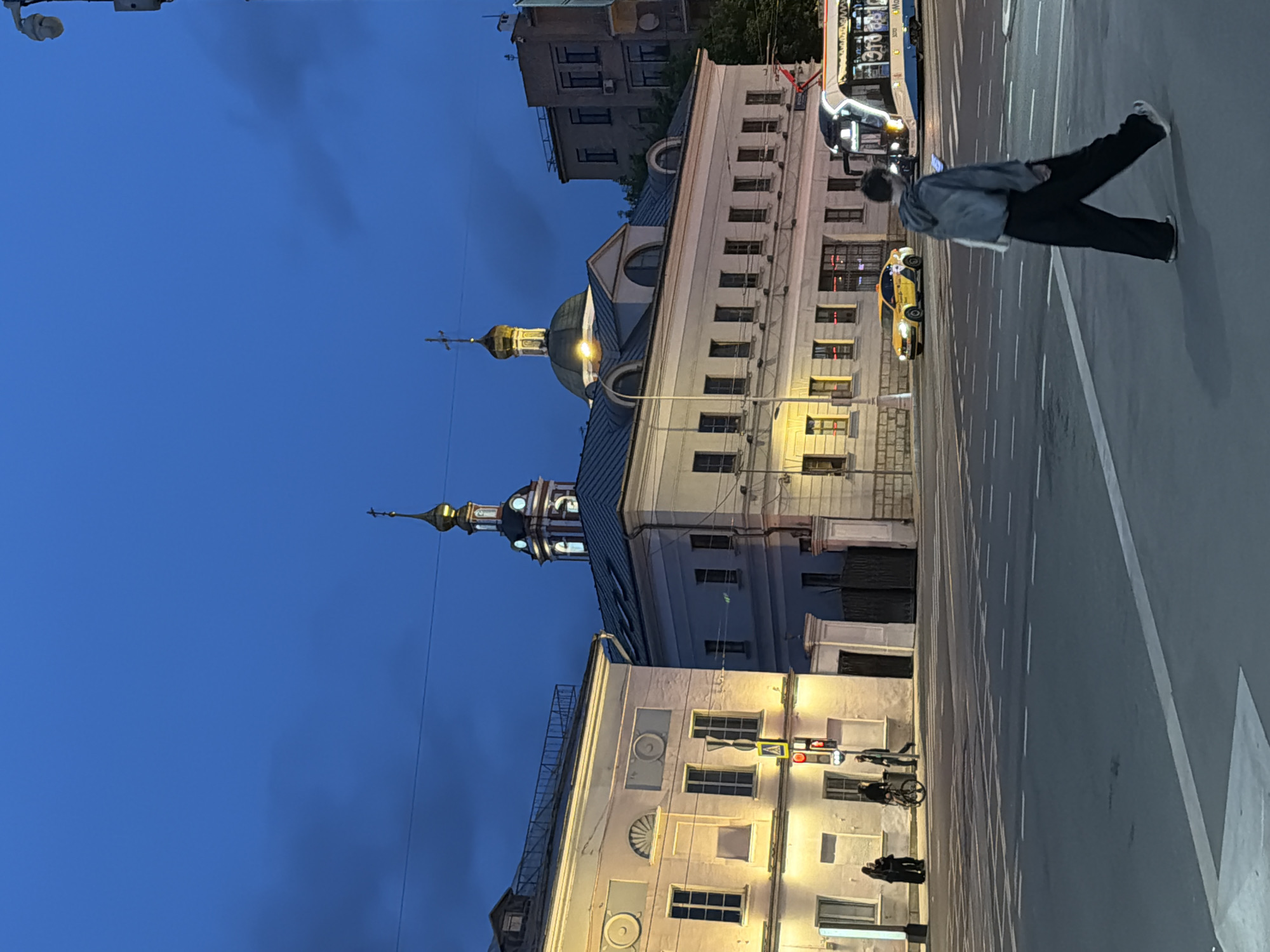The image depicts a street scene at dusk in Moscow, Russia. Several buildings line the street, including a prominent building with a dome and spire, likely a church or cathedral, illuminated by streetlights. A person is walking on the sidewalk, and other people are standing nearby. The street is lined with white lines, indicating lanes, and a yellow taxi is visible. A bus is stopped on the street. The sky is a deep blue color, indicating either early evening or late twilight. Traffic signals are visible.