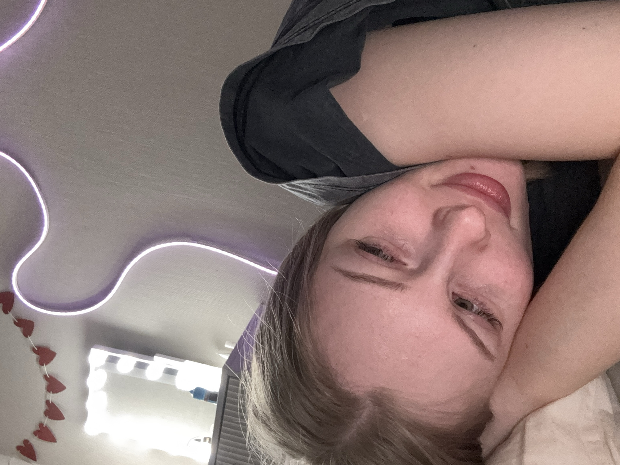 A young fair-skinned person with light brown hair is lying down, possibly in bed, with their head resting on what appears to be a pillow or soft surface. They are wearing a dark-colored t-shirt. A portion of their arm is visible above their head. The person is looking towards the camera.
In the background, there is a white ceiling with a curved purple LED light strip. A vanity mirror with lights and a string of red heart decorations are also visible in the background.
There is no information in the image to indicate the location is Perm, Russia.