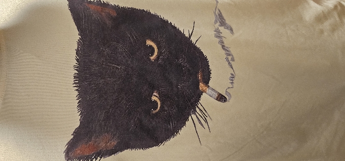 The image presents a close-up, vertical view of a light beige or pale yellow textile surface, likely a piece of clothing, featuring a graphic print. The central subject is a stylized illustration of a black cat's head, occupying the upper portion of the frame. The cat has dark, textured black fur, distinct golden-yellow eyes with horizontal pupils, and reddish-brown inner ear details. Several thin black whiskers extend from its muzzle. A lit cigarette, showing a white filter and an orange-brown burning tip, is depicted held horizontally in the cat's mouth. Wisps of gray smoke are illustrated rising from the cigarette to the right. The textile background is uniform in color and exhibits a faint woven texture. No people, overt actions, interactions, or specific environmental details beyond the fabric texture are present. The image does not provide any visual information to determine a city location in Kamenz - Kamjenc, Germany.