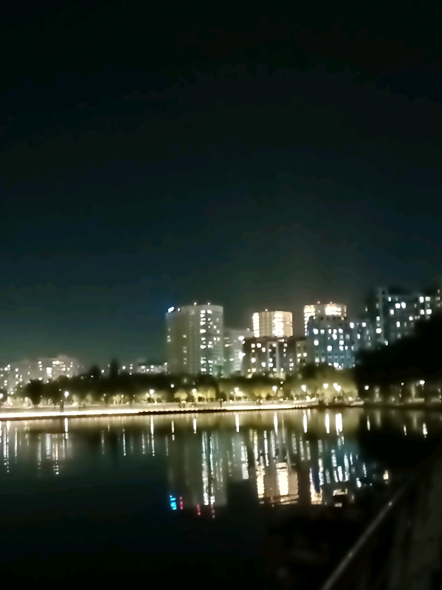 The image shows a nighttime city skyline with buildings and lights reflected in a body of water. The buildings are tall and modern, with many windows illuminated. The water is dark and still, with the lights from the buildings creating a shimmering reflection. There are also some trees visible along the waterfront, which are silhouetted against the lights. The sky is dark and overcast, with no stars visible. The overall impression is one of a peaceful and serene urban landscape.