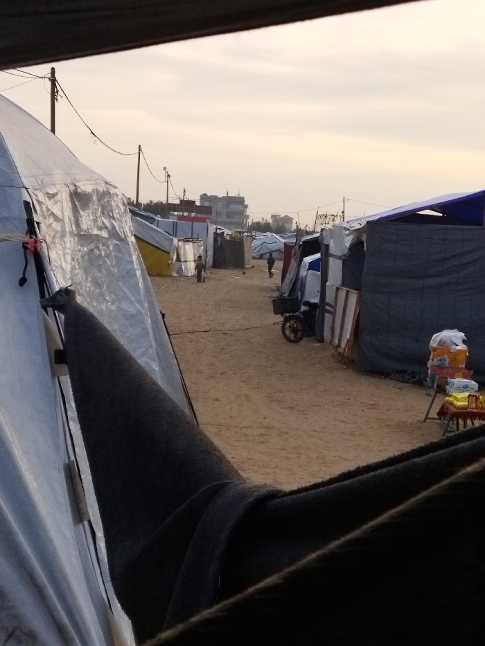 The image displays a makeshift settlement in Khan Yunis, Palestinian Territory, viewed from inside a tent-like structure. A sandy, unpaved path stretches into the midground, flanked on both sides by numerous temporary shelters constructed from white, blue, dark grey, and yellow tarpaulins, as well as other salvaged materials. Utility poles with overhead wires are visible along the left side of the path. A small, dark-skinned child walks alone on the left side of the path, with another adult figure further in the distance, both moving away from the camera. A small motorcycle or tricycle is parked on the right side of the path next to a shelter. On the far right, a small red table functions as a makeshift stall, displaying packaged goods and items in white plastic bags. Taller, more permanent buildings are discernible in the distant background under an overcast sky. The foreground is framed by dark fabric and a light-colored tarp.