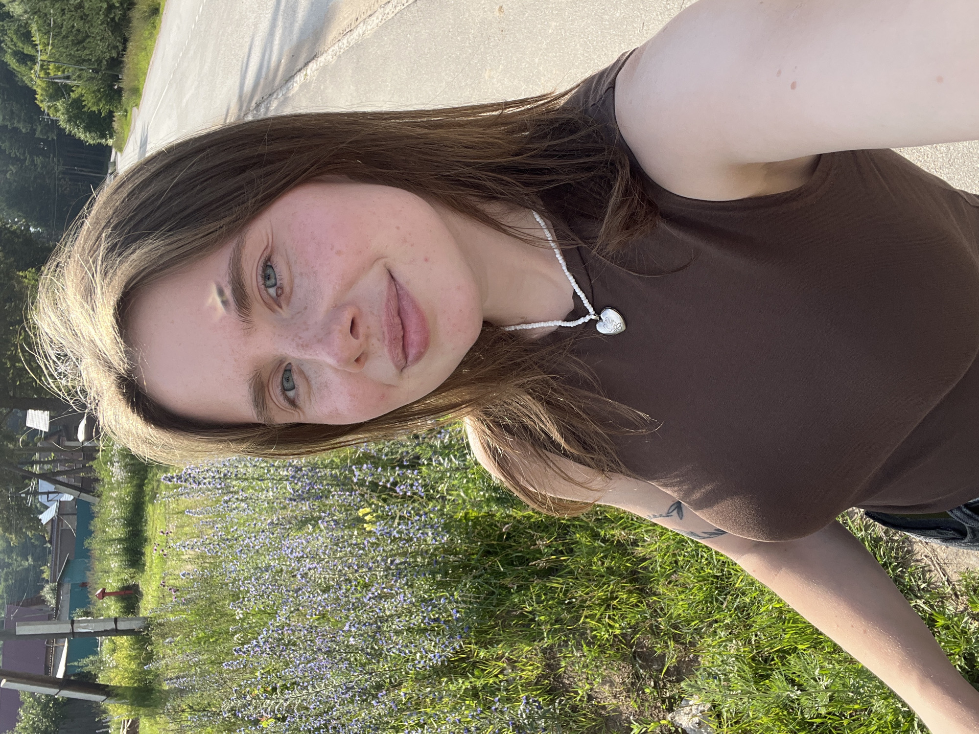 A young woman with light brown hair and blue eyes is taking a selfie outdoors. She is wearing a brown sleeveless top and a white beaded necklace with a heart-shaped pendant. Her left arm, visible on the left side of the image, has a tattoo. She is standing next to a field of purple flowers and tall green grass. In the background, there is a road, trees, and some buildings that appear to be in a rural or suburban setting. The image is well-lit, suggesting it was taken during the daytime with sunlight casting shadows. The environment suggests a warm season. The specific location in Chaikovsky, Russia is not determinable from the visual information alone.