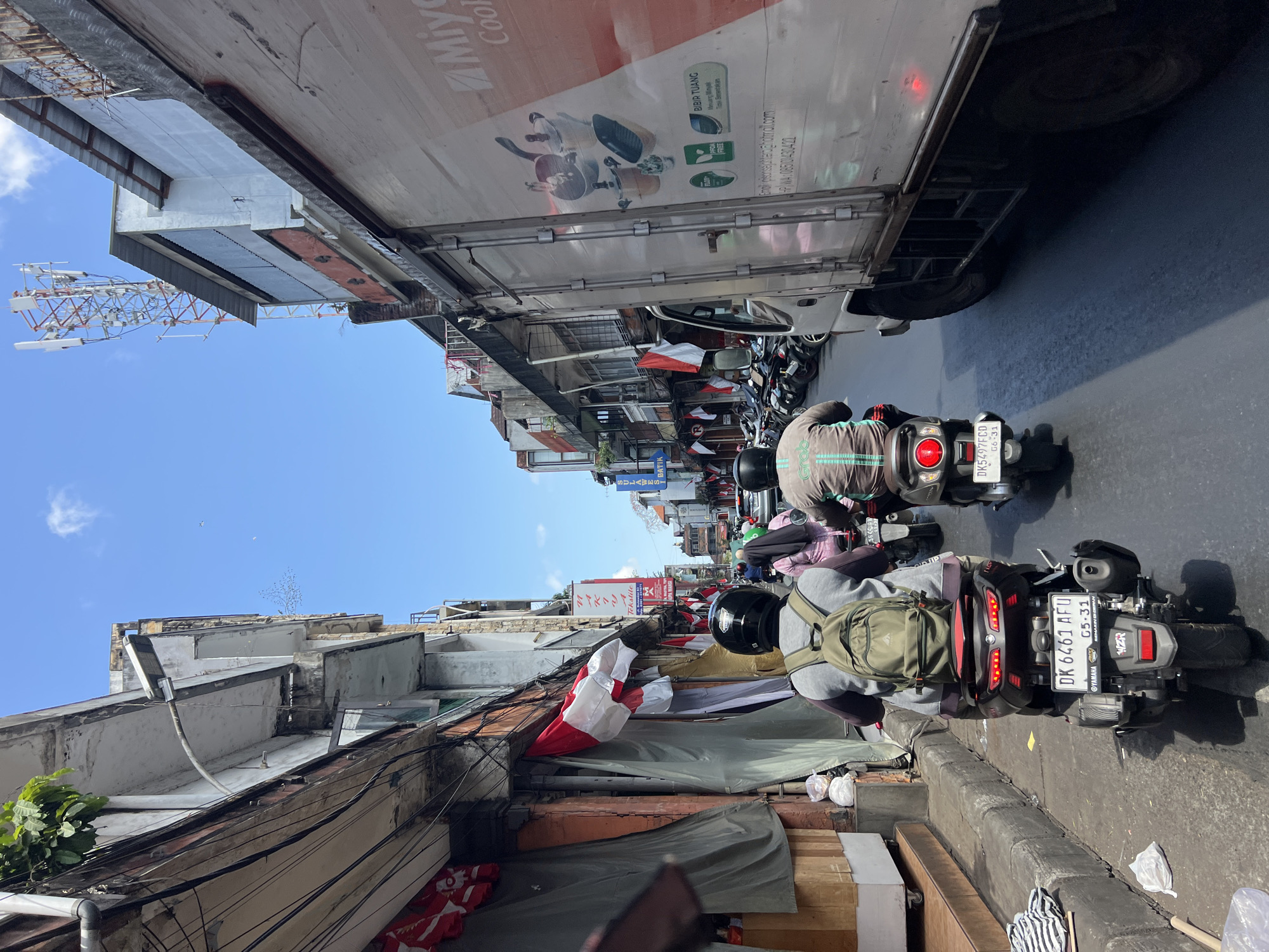 In Denpasar, Indonesia, a street scene unfolds under a bright blue sky with scattered clouds. The narrow road is lined with multi-story buildings, some displaying signs for textiles and batik. Indonesian flags, red and white, are draped along the facades of some structures, suggesting a festive or commemorative occasion. A large white truck with the "Miyako" logo, featuring images of cookware, is prominent on the right side of the frame, partially obscuring the buildings. The street is busy with traffic, primarily motorcycles. In the foreground, two individuals are riding motorcycles. The rider in the immediate foreground, wearing a black helmet and a grey jacket with a light green "Grab" logo on the back, has a green backpack. Their motorcycle's license plate reads "DK 6461 AFU". To their right, another motorcycle rider, wearing a pink headscarf and pink shirt, is partially visible. Further down the street, other motorcycles and a vehicle can be seen, indicating moderate urban traffic. The buildings appear to be a mix of commercial establishments and residences, with some showing signs of wear and tear. Electrical wires are visible overhead, strung across the street between buildings. The ground is paved with asphalt, and a concrete curb runs along the side of the road.