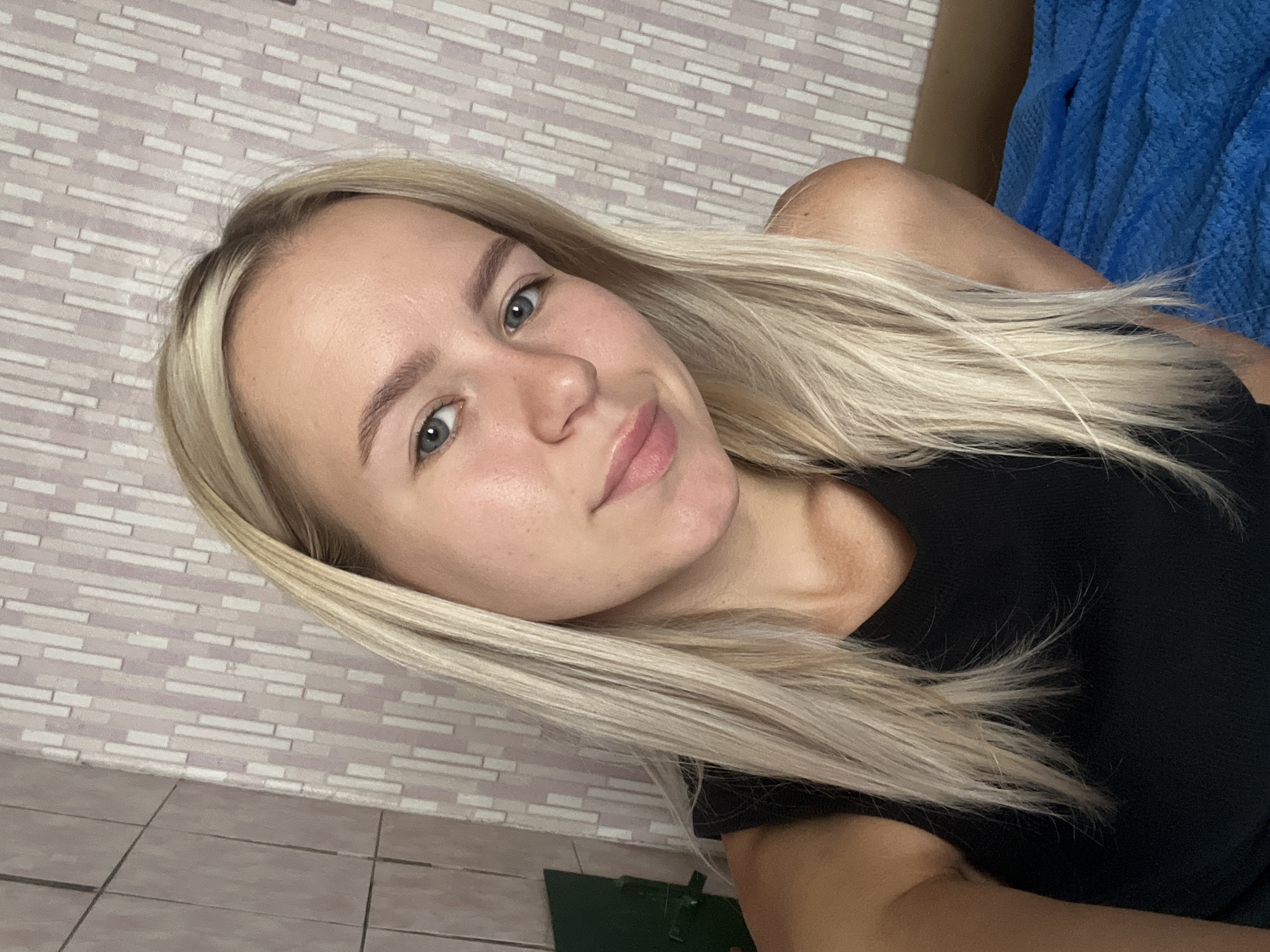 A woman with blonde hair and blue eyes is taking a selfie. She is wearing a black top. The background is a wall with a pink and white striped wallpaper. There is also a glimpse of a green object on the left side of the image and a blue blanket on the right. The setting appears to be indoors, likely a room.