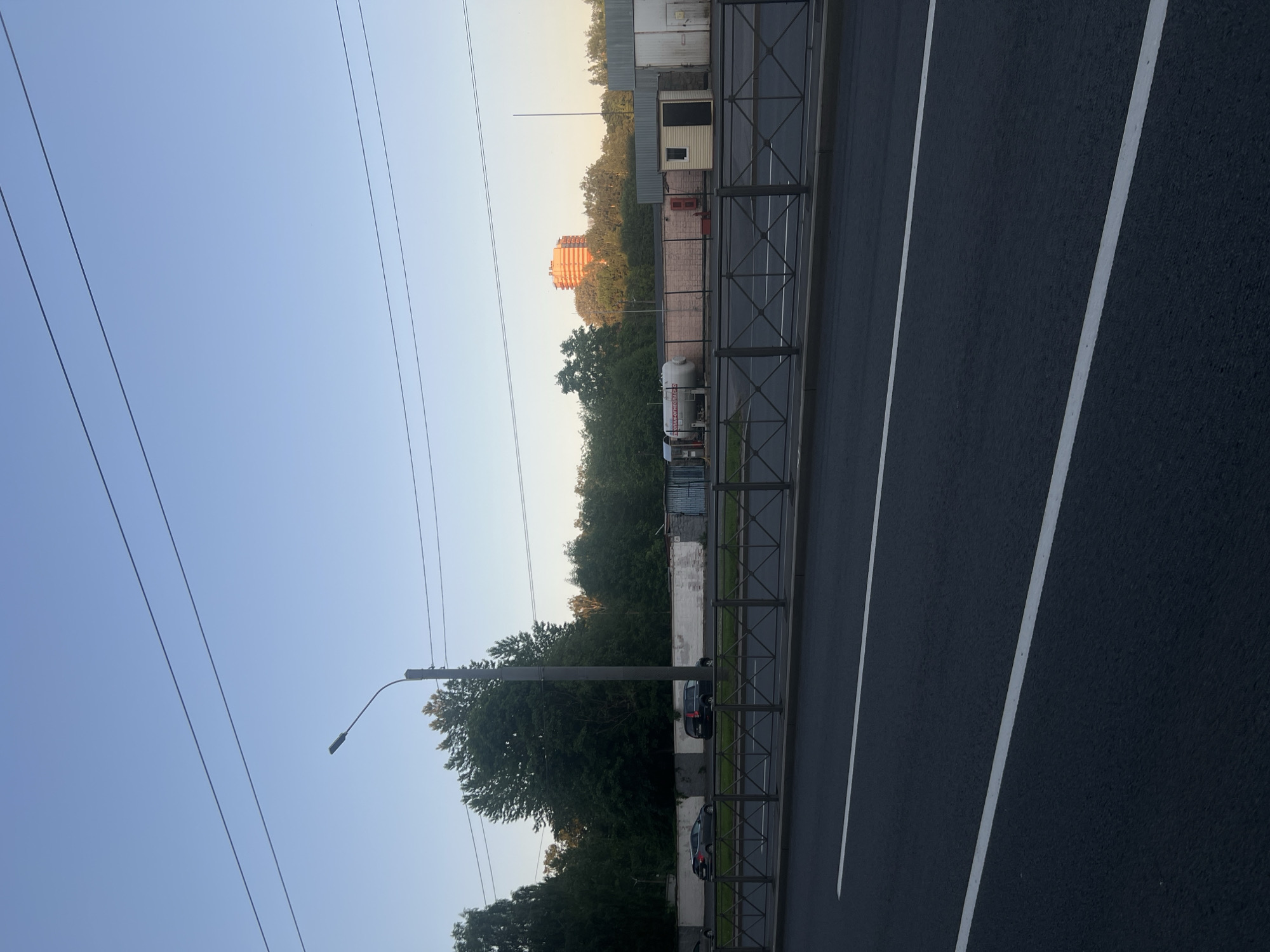 The image depicts an outdoor urban scene at an unspecified time, likely late afternoon or early morning, under a clear, light blue sky in the city of Unknown, Russia. Overhead, two prominent parallel power lines traverse the upper portion of the frame horizontally, with a third lower line visible. A dark grey street light pole, featuring a curved arm and a modern, rectangular LED fixture, stands on the left side, with its light off.
In the background, dense green deciduous trees form a prominent treeline, partially obscuring the landscape behind. Above the trees on the far right, a multi-story, orange-colored building is visible, illuminated by low-angle sunlight. Below the treeline, a long, white, weathered wall runs horizontally, separating the roadside from various properties. Behind this wall on the left, two cars are parked parallel: a dark grey sedan and a dark SUV. Further right, behind a lower, light pinkish-brown brick wall, an industrial or commercial area is visible, featuring a large white horizontal gas storage tank with red Russian text, including "ОГНЕОПАСНО" (FIRE DANGEROUS). Adjacent to the tank are several single-story structures, including a small, light beige building with a dark door and window, and a larger grey building with a garage-style door.
The midground features a dark metal fence with a diamond-patterned grid running horizontally across the entire width of the image, separating the roadside from the vehicular traffic. A narrow strip of green vegetation is present between the fence and the road, bordered by a dark grey curb. The foreground is dominated by a dark asphalt road with two distinct, solid white lines running parallel across its width, indicating traffic lanes. No individuals are visible, and no active movements or interactions are depicted.