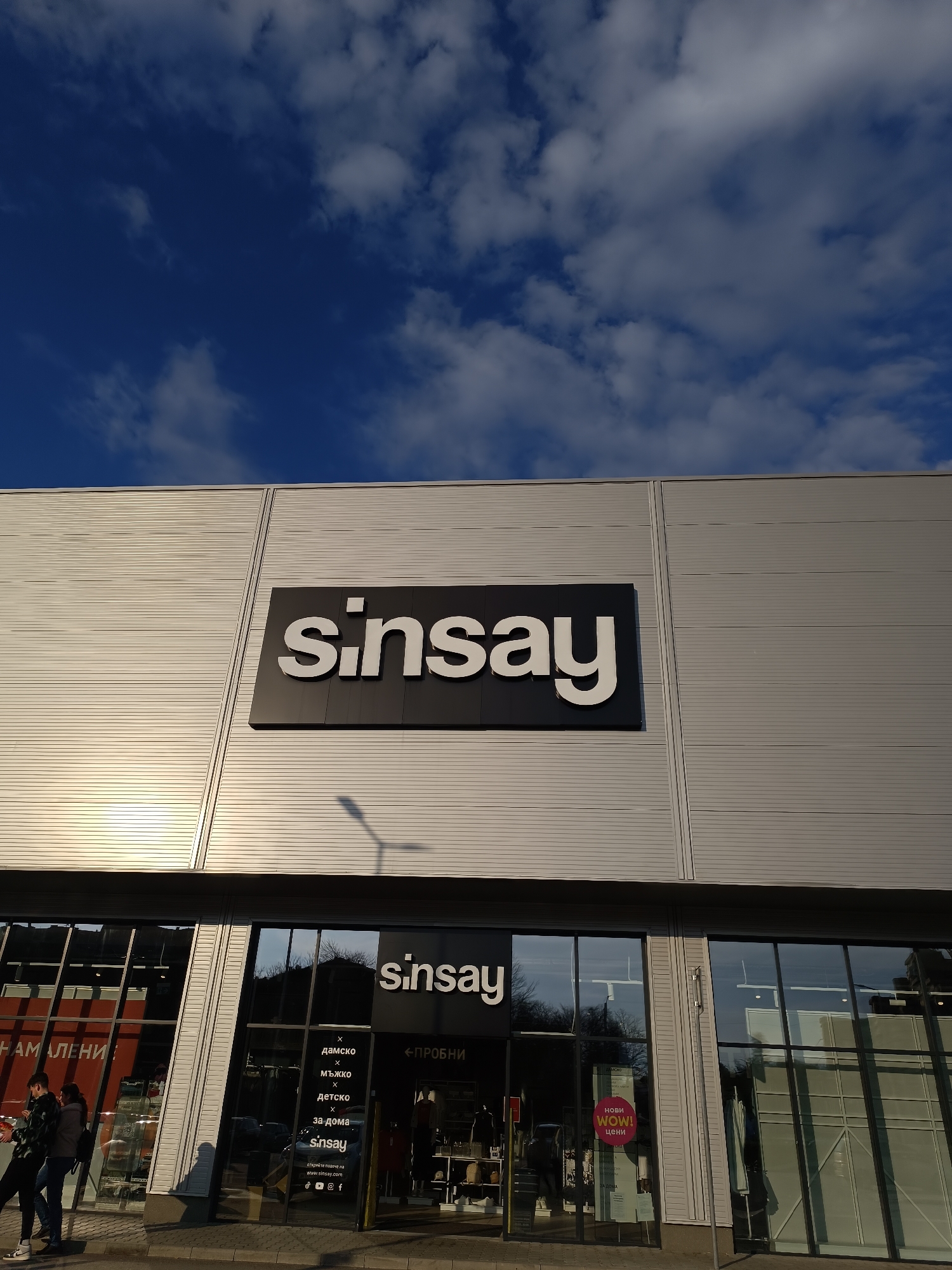 Here is a detailed description of the image:
The image shows the exterior of a "Sinsay" clothing store in the city of Pleven, Bulgaria. The store is a large, boxy building with a light grey, horizontally-paneled exterior. There is a large "Sinsay" sign with white letters on a black background mounted near the top of the building's facade.
Below the large sign, the store's entrance is visible, featuring glass windows and doors. Above the entrance, there is another smaller "Sinsay" sign. On the glass windows, the words "дамско" (women's), "мъжко" (men's), "Детско" (children's), and "за дома" (for the home) can be seen, indicating the store's offerings. A sign with "ПРОБНИ" (fitting) and an arrow points the direction. There's also a round pink sign that says "нови WOW! цени" (new WOW! prices).
On the left side of the image, two people are visible walking near the store. The sky is blue with scattered clouds. A lamppost is seen casting a shadow on the building. The setting is likely daytime, given the lighting and presence of people.