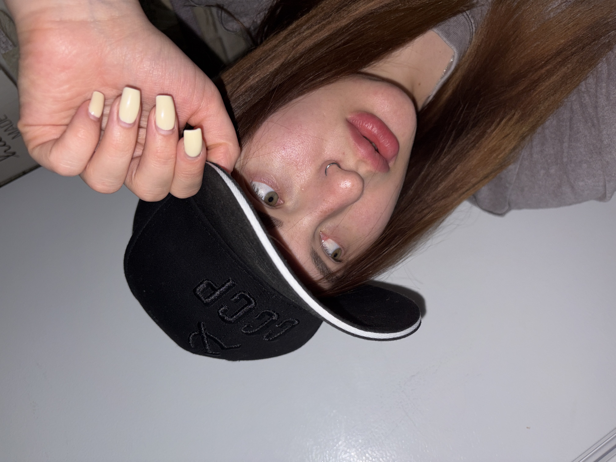 A woman with light skin and brown hair, wearing a grey shirt, is lying on a light surface, possibly a bed or table. She is wearing a black baseball cap with white trim and Cyrillic lettering. Her nails are painted a pale yellow color. She has a nose ring and is looking at the camera with her mouth slightly open. The location is not identifiable as being in the city of Port-Katon, Russia.