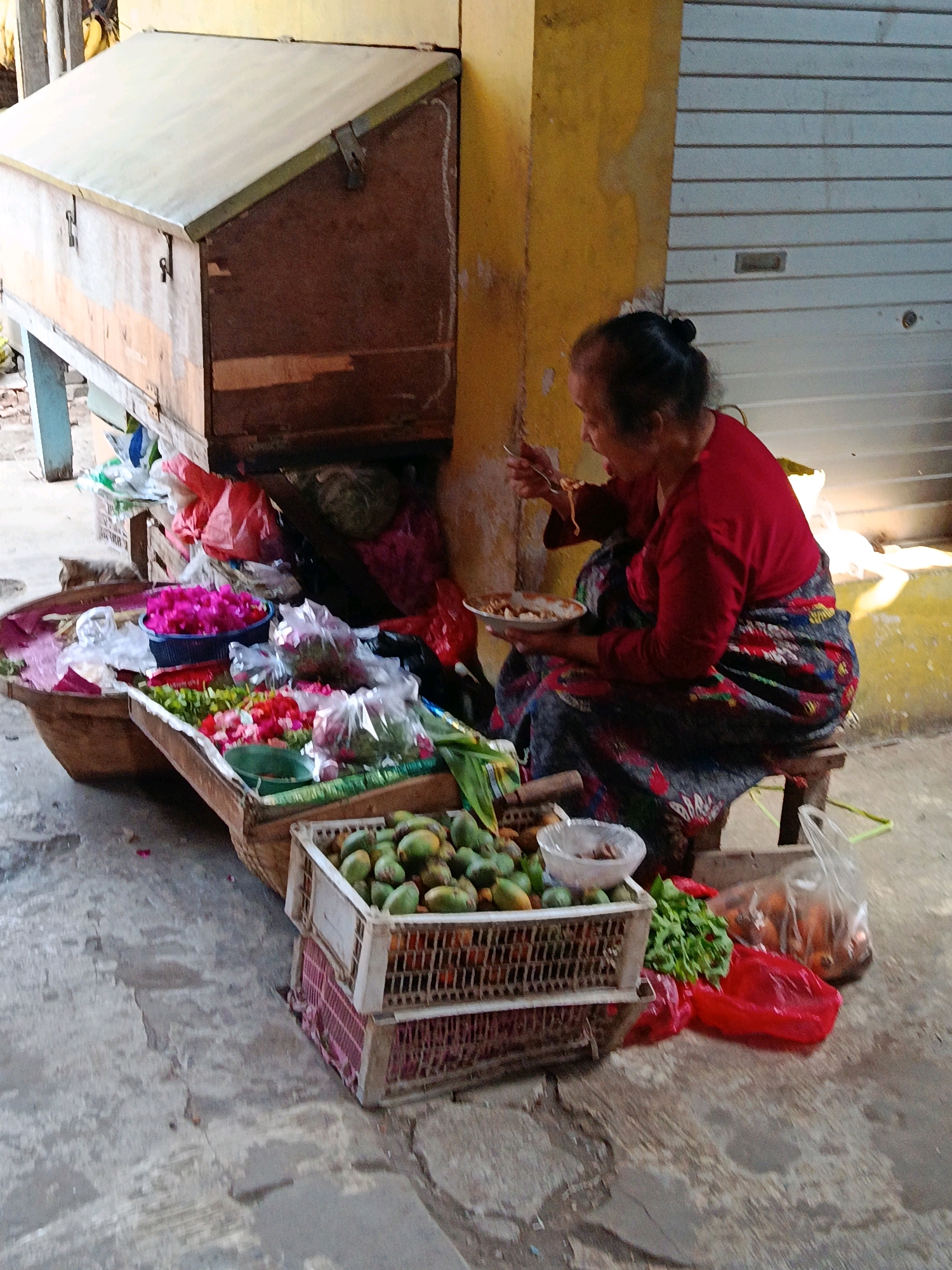 An elderly woman, likely of Indonesian descent, is seated on a low wooden stool and eating from a plate with a fork. She is wearing a red long-sleeved shirt and a vibrantly patterned sarong. She is positioned next to a market stall laden with various fruits and vegetables. In front of her, there are two stacked white plastic crates filled with green produce, possibly unripe mangoes or similar fruits. To the left of these crates, a large woven basket overflows with pink and red flowers. Further back, several plastic bags, some red and some transparent, contain more produce and groceries. The stall itself is constructed of wood, with a slanted metal lid on the left side. The woman is facing a yellow wall, and to her right, a silver, corrugated metal roll-up door is visible. The ground is paved with concrete, showing signs of wear and tear. The overall impression is a snapshot of daily commerce at a local market, possibly in Mlawang, Indonesia, given the context.