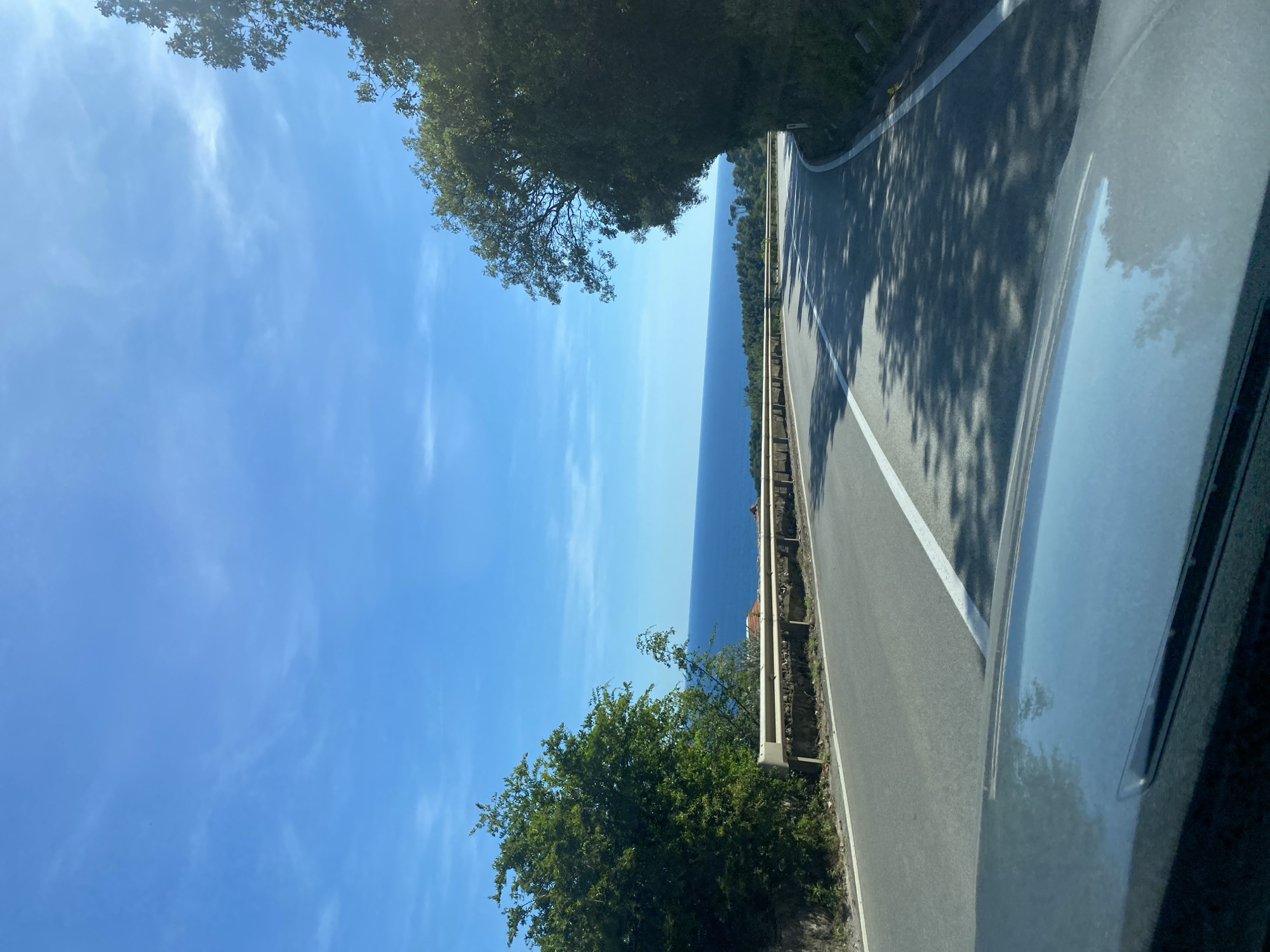 The image captures a scenic coastal road in Nerin, Montenegro. A two-lane asphalt road curves to the right, bordered by a metal guardrail on the left and a steep, tree-covered incline on the right. The sea, a deep blue, stretches out to the horizon under a clear, bright blue sky with scattered wispy clouds. Lush green trees flank the road, with some casting shadows across the asphalt. The foreground shows the hood of a car, reflecting the sky and trees, suggesting the photo was taken from inside a vehicle. In the distance, glimpses of red-tiled roofs can be seen peeking through the trees on the left side of the road.