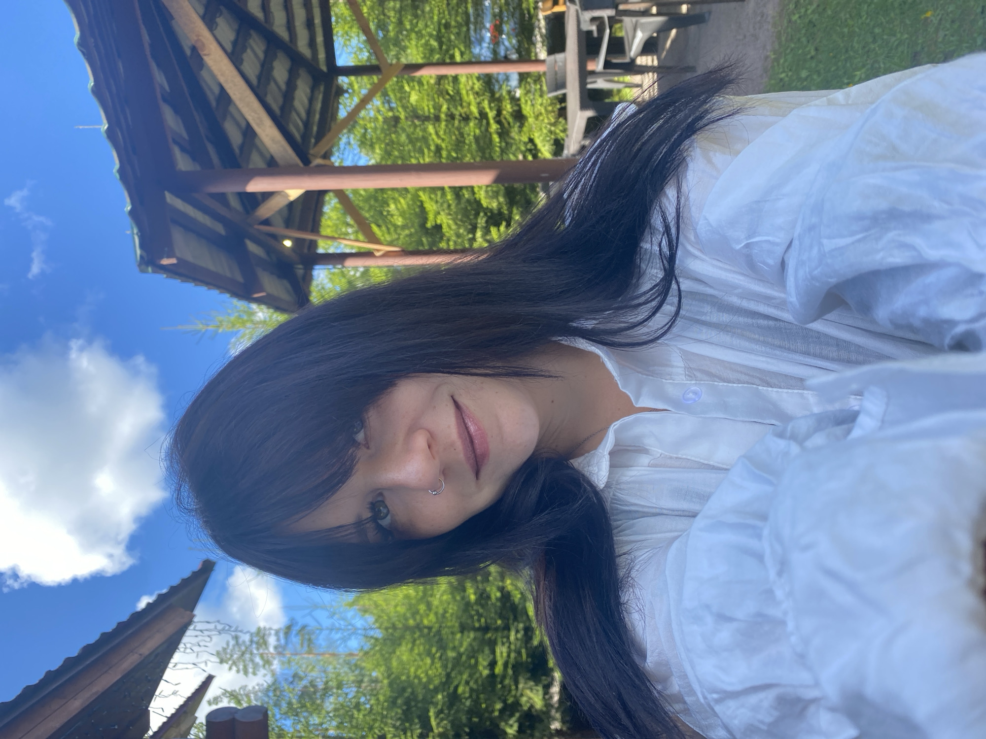 A young woman with long, dark hair and bangs is taking a selfie outdoors on a sunny day. She is wearing a white, button-up shirt. She has a nose ring. In the background, there is a wooden gazebo, trees, and a blue sky with white clouds. The location is the city of Krasnoflotskoe, Russia.