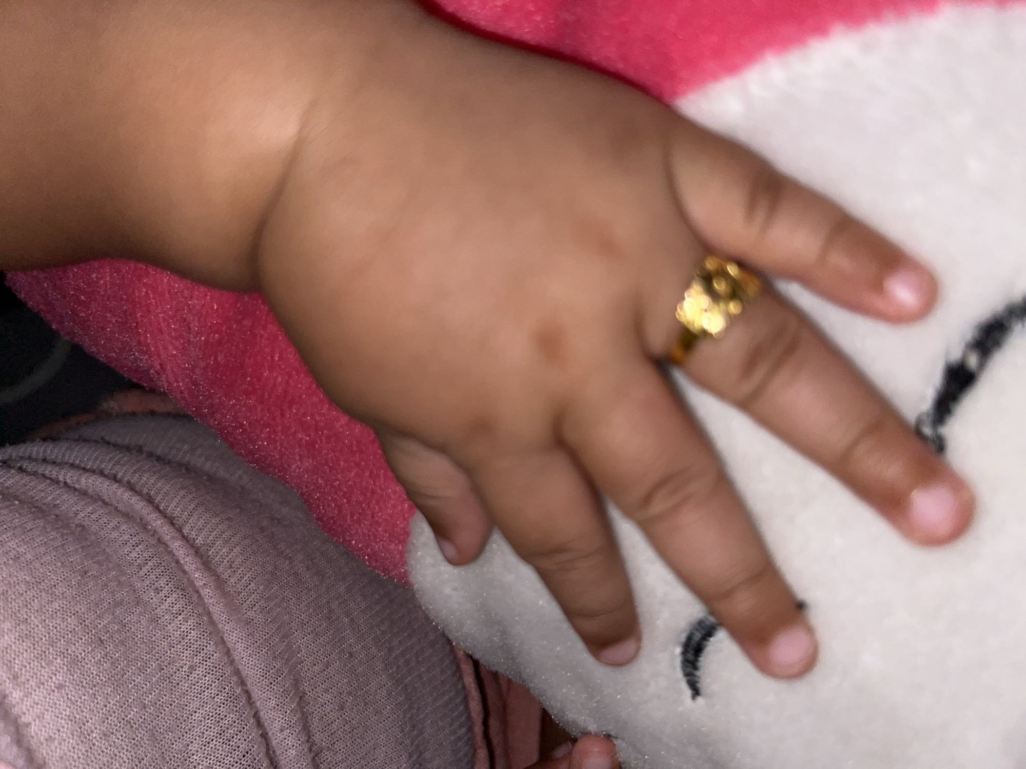 A baby's hand with a gold ring on the ring finger rests on a pink and white surface. The hand is in the foreground. Part of a pink fabric and a light purple garment are also visible. There is no visible information in the image that would indicate the location of the photo is in Lhokseumawe, Indonesia.