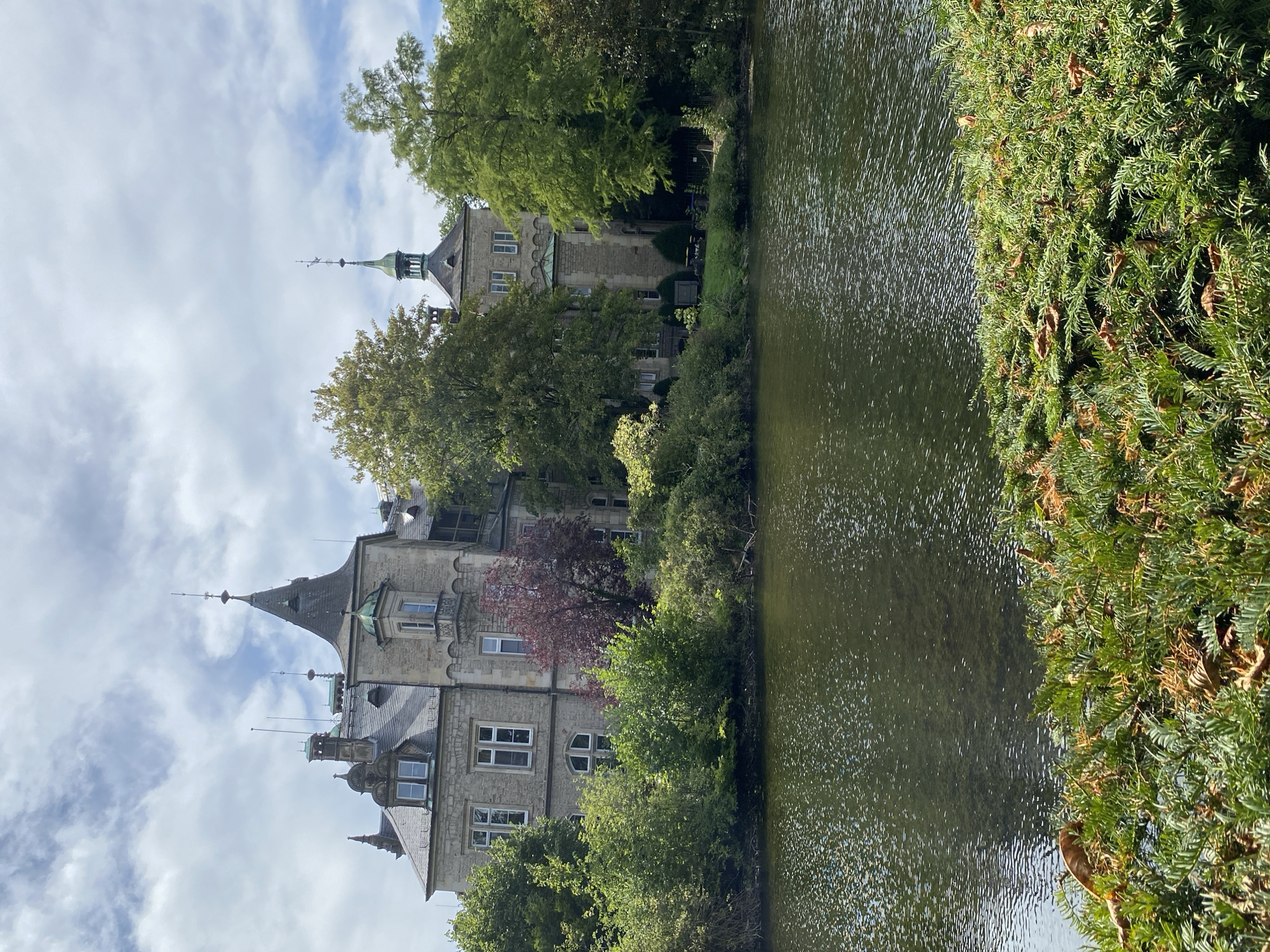 The image depicts the Bückeburg Castle (Schloss Bückeburg) located in Bückeburg, Germany. The castle, a prominent historical landmark, is situated on the bank of a calm body of water, likely a pond or moat. The architecture of the castle is a blend of Baroque and Renaissance styles, characterized by its stone facade, multiple turrets topped with green spires, ornate windows, and a steep, grey roof. Lush green trees and dense foliage surround the castle, with some trees appearing to be deciduous and showing hints of autumn colors. The water's surface reflects the sky and surrounding greenery, creating a shimmering effect. In the foreground, a thick hedge with some dry leaves adds texture to the scene. The sky is partly cloudy with scattered white clouds against a pale blue background. There are no people visible in the image.