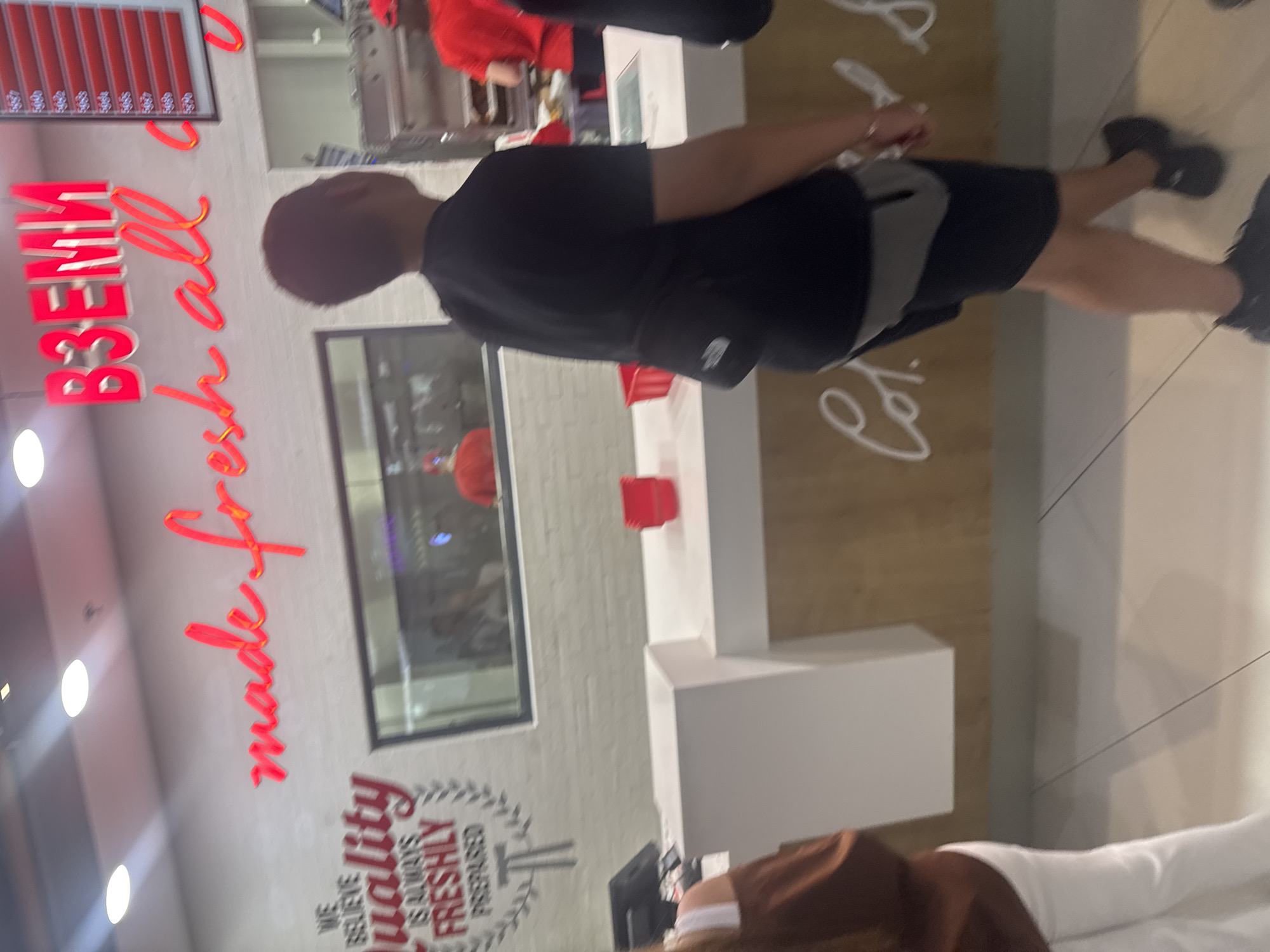 A person in a black shirt and black shorts with a grey fanny pack is standing at a counter in what appears to be a fast food restaurant. In the background, a sign with the phrase "Вземи made fresh all..." is visible. Another sign below it states, "We believe quality is always freshly prepared." The location is likely in Plovdiv, Bulgaria based on the Cyrillic letters of the word "Вземи". Other people are present at the counter and behind it in the kitchen area, and a person in brown clothes and white pants is partially visible at the bottom of the image. The floor is tiled and light colored.