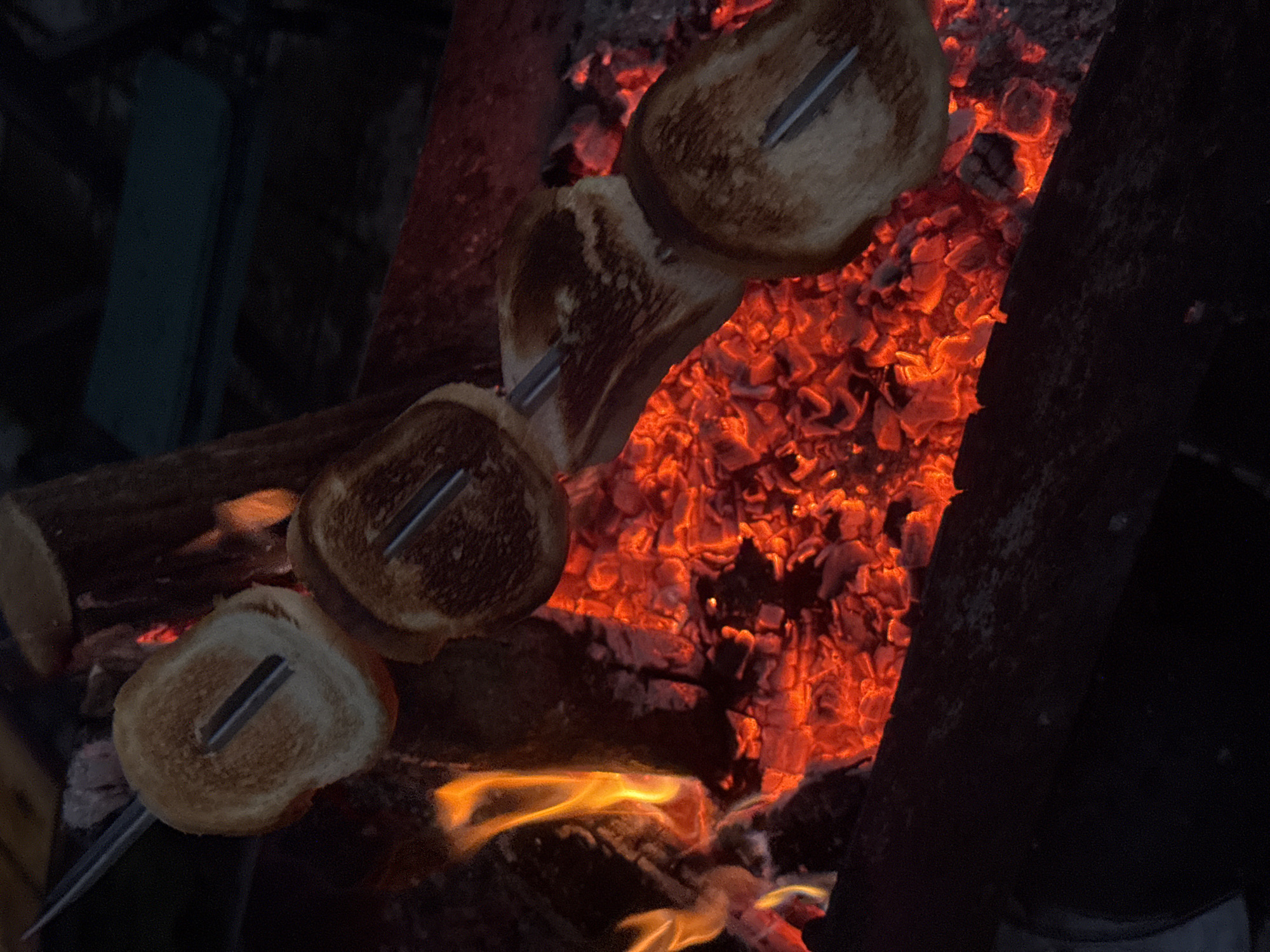 In the dark, rural setting of Автомобилист-1 Снт, Russia, a campfire blazes with bright orange flames and glowing red embers. On a metal skewer, four slices of toasted bread are being roasted over the heat. The bread slices are golden brown, some with darker toasted spots, indicating they are being cooked. The skewers are angled downwards towards the coals, suggesting the bread is in the process of toasting. The surrounding environment is dimly lit, with the fire being the primary light source. The composition is a close-up view of the campfire and the food being prepared, emphasizing the act of roasting.