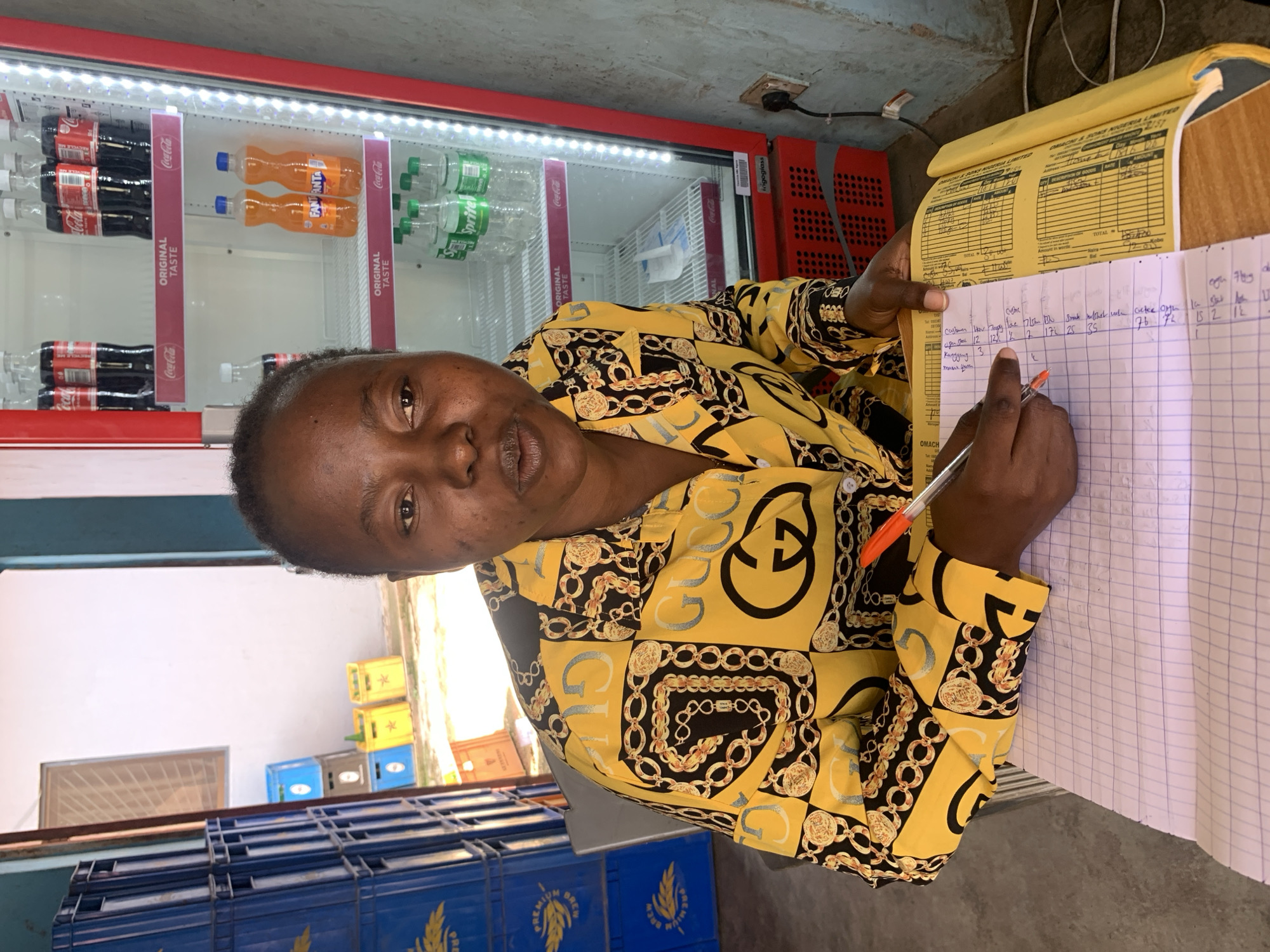 A woman is sitting at a wooden desk in what appears to be a small shop or convenience store in Jos, Nigeria. She is wearing a yellow and black patterned shirt with gold chain and 'GUCCI' logos. Her left hand is resting on a pink and white checkered notebook, and her right hand is holding a red pen, actively writing in the notebook. The notebook is open to a page with columns of handwritten entries in a dark ink.
Behind her, to her right, is a red and white beverage cooler with internal lighting. The cooler is stocked with several bottles of Coca-Cola, Fanta, and Sprite. To her left, a stack of blue plastic crates, possibly for beverages, is visible, along with some yellow and blue crates stacked further back. The walls of the shop are a light color, and a power outlet is visible on the right wall with a cable connected to the cooler. The overall environment suggests a commercial setting where beverages are sold. The lighting in the scene is artificial, likely from the cooler and ambient shop lighting.