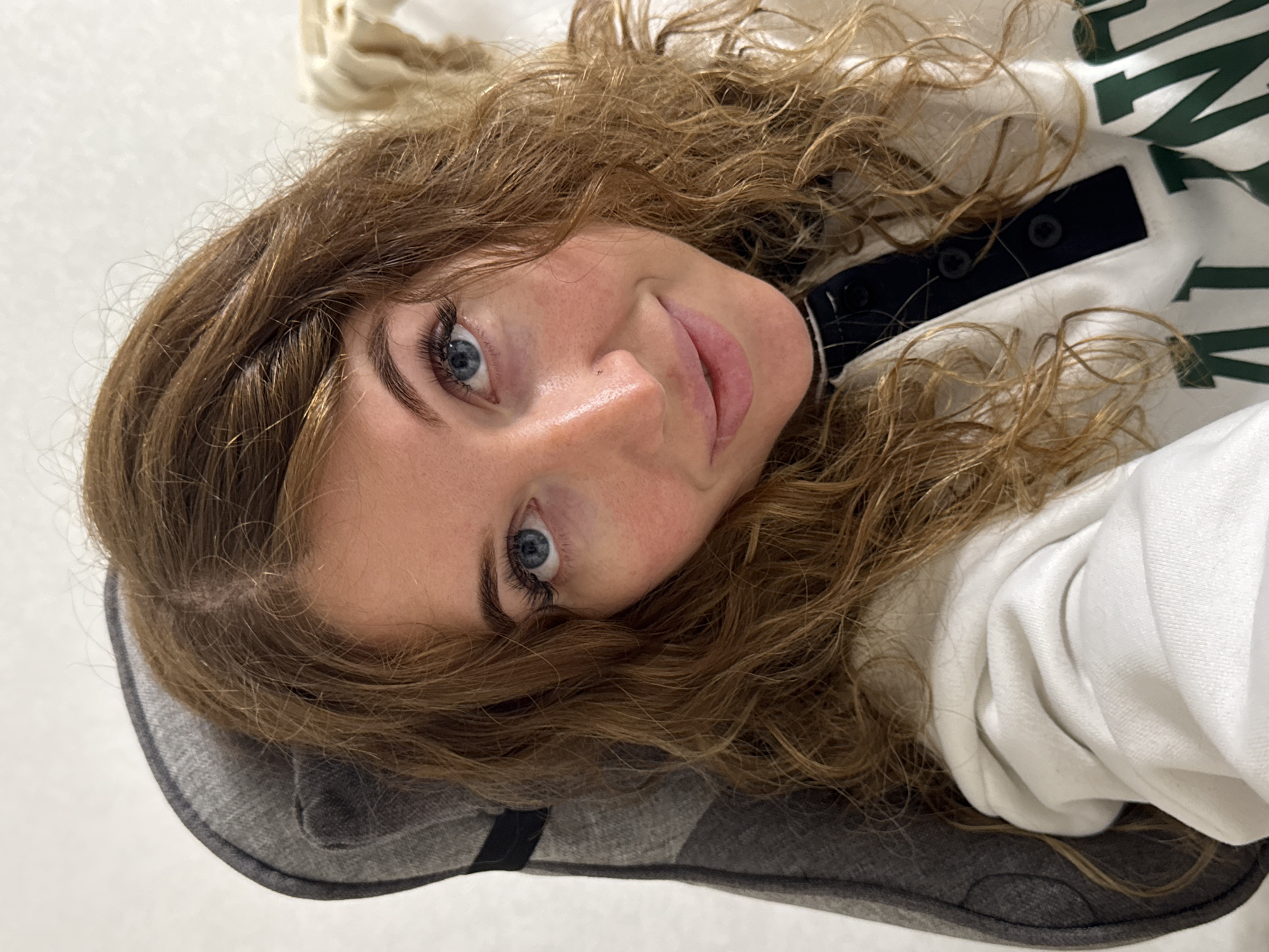 A woman with light brown, curly hair and blue eyes is lying down, her head resting on a gray cushion or pillow. She is wearing a white top with dark green lettering and a dark navy collar with buttons. The background is a light, possibly white, wall. The location is unspecified but suggested as Chaikovsky, Russia.