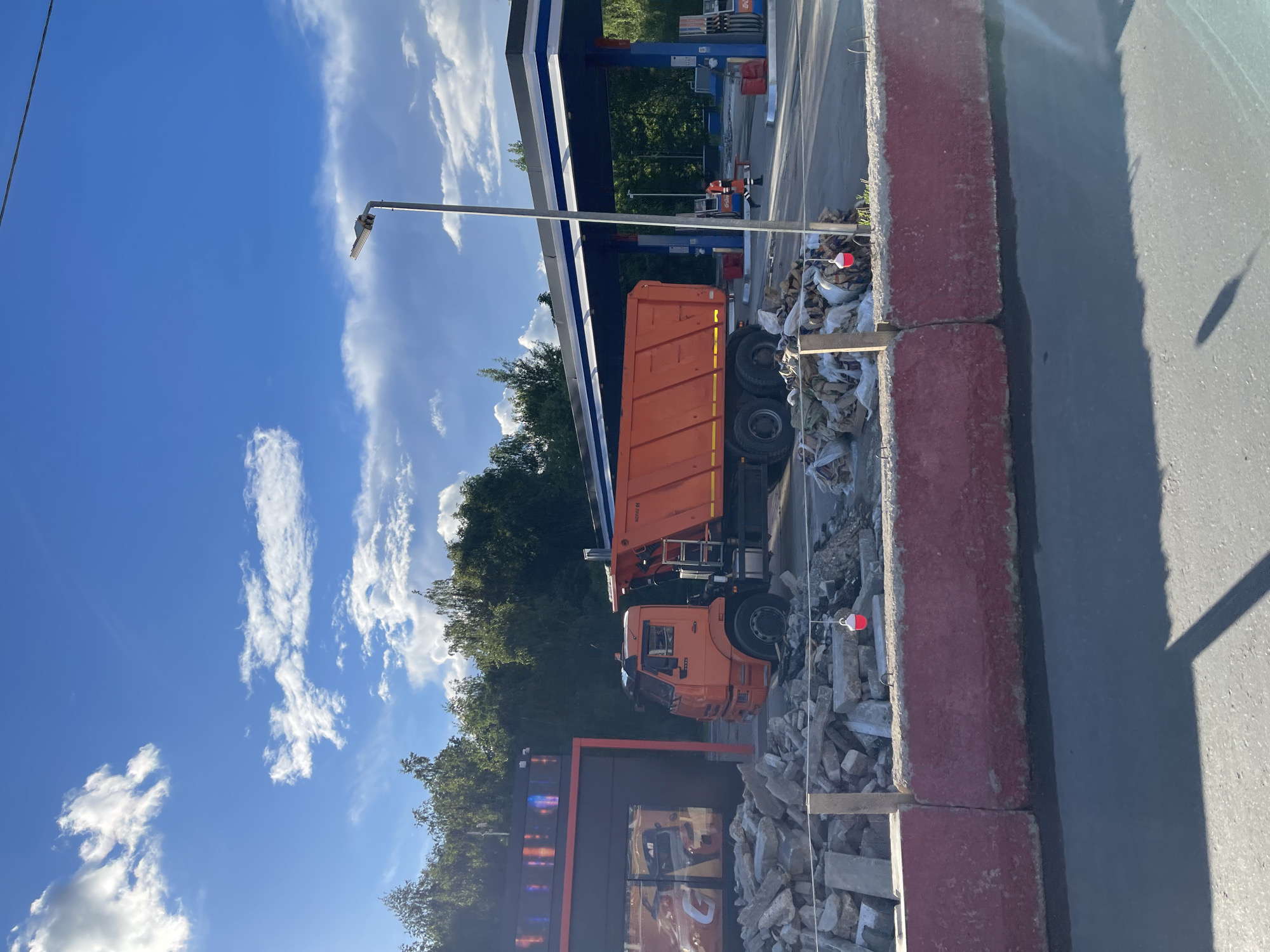 An orange dump truck is parked near a building under construction, likely in Moscow, Russia. The truck is surrounded by debris, including concrete blocks and white bags of construction materials. Behind the truck, there is a gas station with a blue and white canopy. In the background, there is a tree line and a blue sky with scattered clouds. The scene is illuminated by bright sunlight.