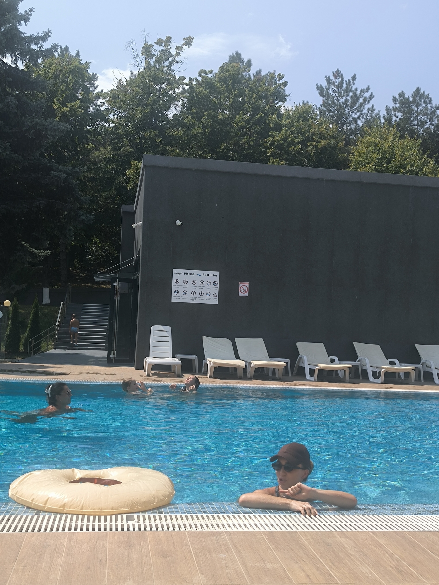 In Ustia, Moldova, a poolside scene unfolds under a clear blue sky with scattered clouds. A dark, modern building with a black facade stands to the right, featuring signs with pictograms indicating pool rules. Adjacent to the building, a set of outdoor stairs leads up to an entrance, with a young person in swim trunks ascending. A large, inflatable ring-shaped float rests on the surface of the bright blue swimming pool. In the foreground, a woman wearing sunglasses and a baseball cap leans on the edge of the pool, her head and shoulders visible above the water. Further in the pool, a woman with her hair tied up swims, and two children are visible, one appearing to splash the other. Several white lounge chairs are arranged on the tiled deck behind the pool. The background is dominated by lush green trees, suggesting a natural or park-like setting surrounding the pool facility.