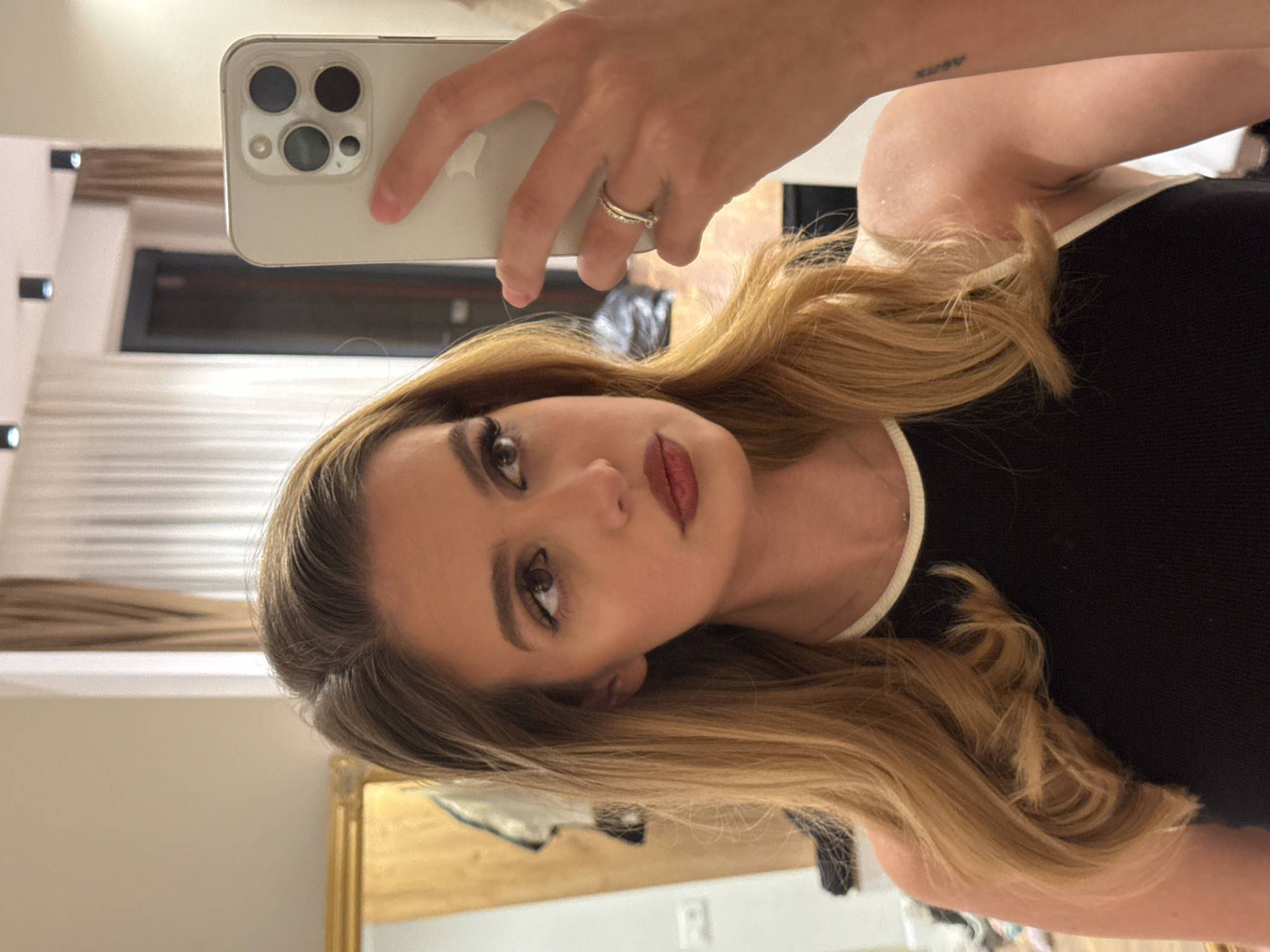 The image is a selfie of a young woman, likely taken in a mirror, as her phone is visible in her hand, pointed towards herself. The image is rotated 90 degrees counter-clockwise.
The subject is a woman with long, flowing blonde hair styled with a side part and loose waves. She has a made-up face, featuring dark eyeliner/mascara and dark red lipstick, and is looking directly into the camera with a neutral, poised expression. She is wearing a black top with a thin white trim along the neckline. Her right hand, holding a light-colored smartphone with a prominent triple-camera system (identifiable as an iPhone), is visible in the upper part of the frame. A silver ring is worn on her ring finger. A small, faint tattoo, which appears to spell "ARMY" in lowercase, is visible on her right upper arm.
The setting is an indoor space, reflected in what seems to be a mirror. The background shows various elements, including a light-colored wall with horizontal lines (possibly blinds or wooden panels), a dark rectangular object, and a golden-framed item, possibly a mirror or artwork. An electrical outlet is also faintly visible on a light-colored wall. The lighting suggests artificial indoor light. The activity taking place is the woman capturing her own image, likely through a mirror selfie. The time of day and weather are not discernible.