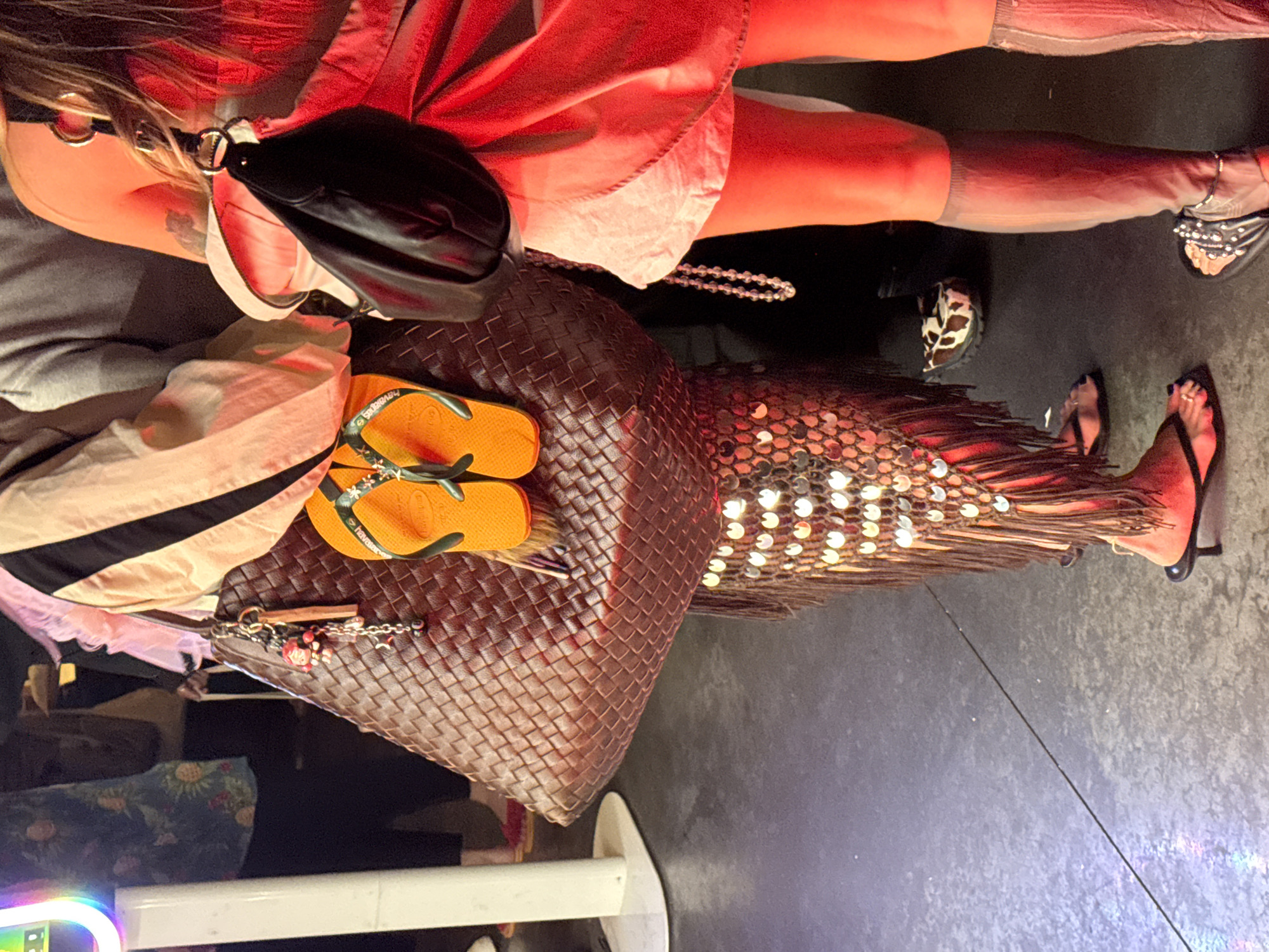 The image shows a close-up view of several individuals in what appears to be an indoor public space. The lighting suggests a low-light environment with some colorful illumination, potentially from a photo booth or decorative lights, as seen on the left side of the frame.
In the foreground, a person is wearing a brown woven tote bag with orange flip-flops dangling from it. The tote bag is large and textured. The person also has a skirt with a fringe and sequin detailing, and is wearing black thong sandals with a thin heel. Another person is partially visible to the right, wearing a white outfit and a black shoulder bag. Their legs are visible, and they are wearing sheer, textured hosiery. The floor is a dark, speckled surface.
In the background, other individuals are visible, including one wearing a grey sweatshirt. Another person in the background is wearing cow-print shoes. The overall scene suggests a social gathering or event. The location is not identifiable from the visual information provided.