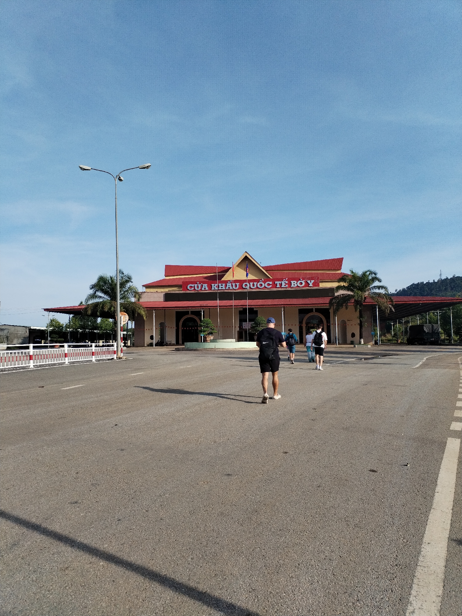 The image depicts a street scene in Bờ Y, Vietnam, at the Cửa Khẩu Quốc Tế Bờ Y border crossing. In the foreground, three people are walking away from the camera towards the border crossing building. The building has a red roof and is flanked by palm trees. A road stretches across the foreground, and a streetlight stands to the left. In the background, there is a truck and a small mountain. The sky is blue.