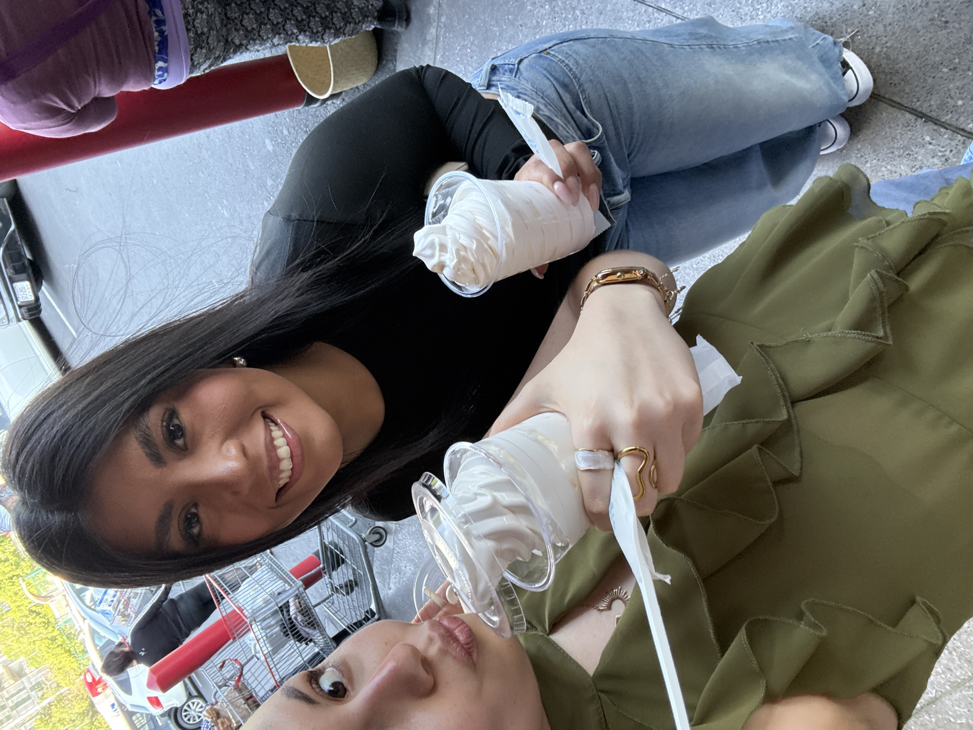 Two women are holding plastic cups filled with ice cream and eating it. One woman has long, dark hair, a black long-sleeved shirt, blue jeans, and white sneakers. The other woman has light skin, a green blouse with ruffles, and gold rings. They are standing near shopping carts, a car, and a road with trees. It seems to be a parking lot in Mexico City, Mexico.